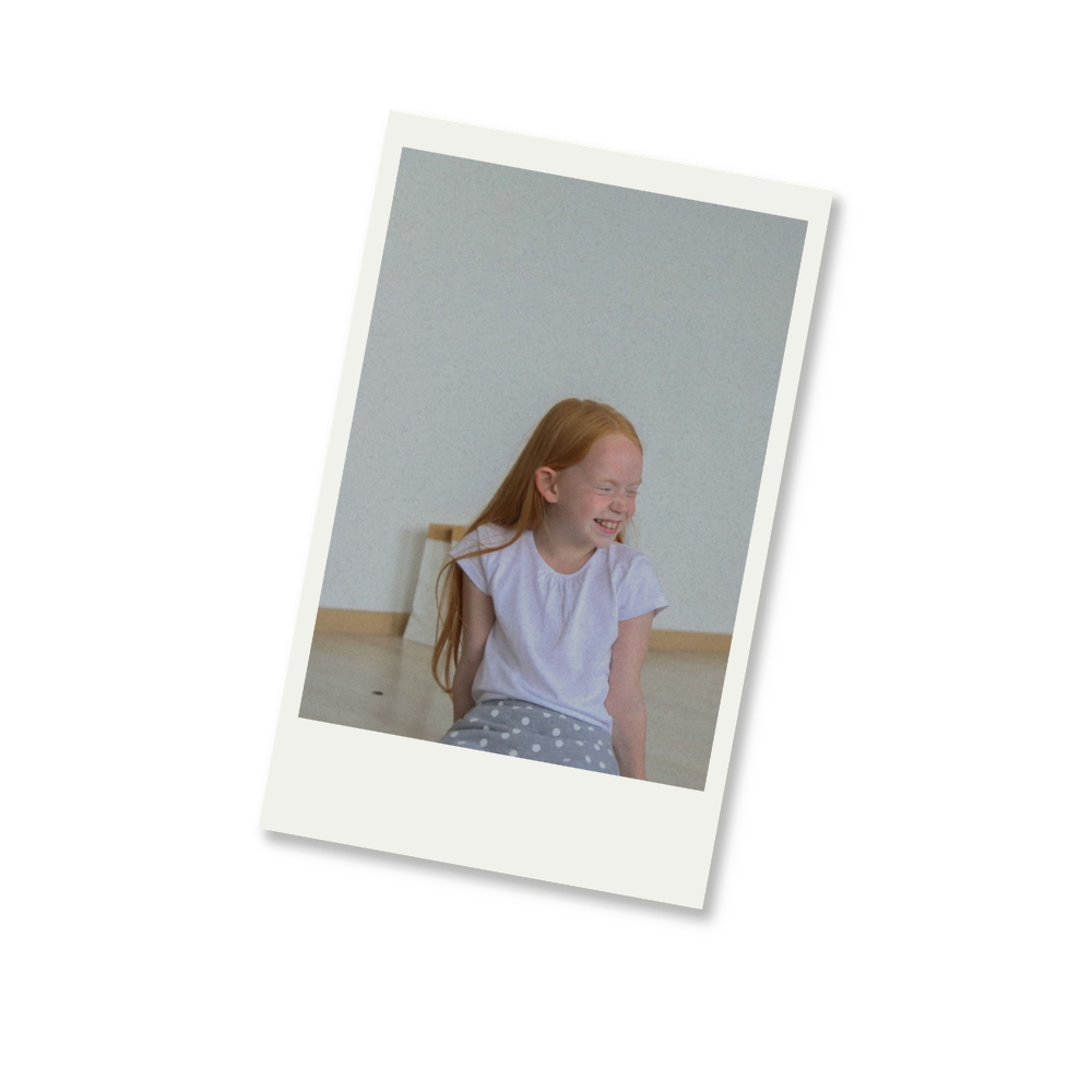 A young girl with red hair laughing and sitting on a wooden floor with a plain light-colored wall behind her.
