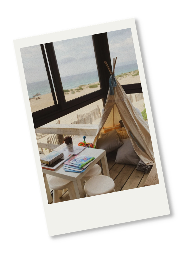 Indoor children's play area near a large window with a view of the beach and ocean, featuring a small table, books, and a fabric teepee tent.