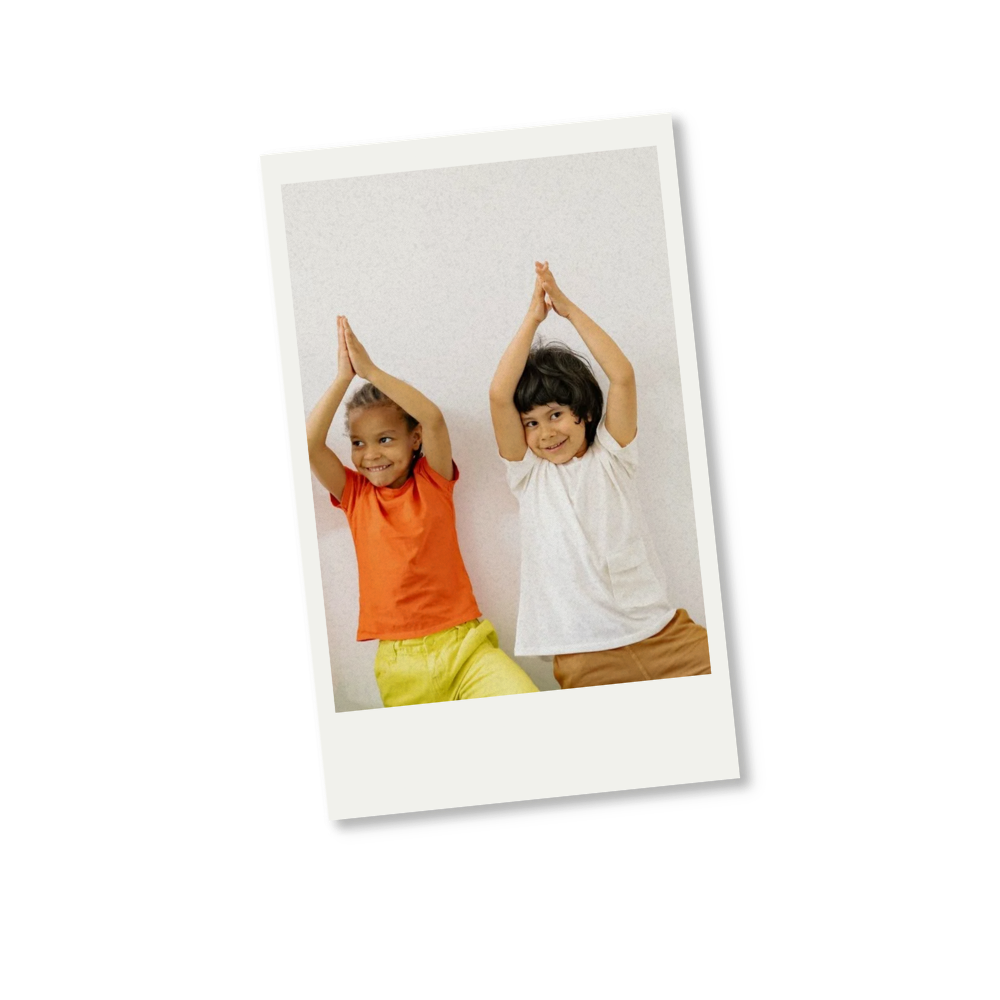 Two children, a girl and a boy, smiling and waving with their hands raised above their heads, standing against a white wall.