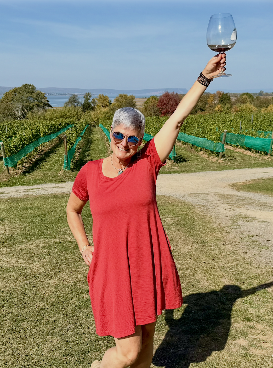 Norma Normington in a red dress and sunglasses smiling and holding a DelBrenna luxion crystal wine glass in a vineyard on a sunny day.