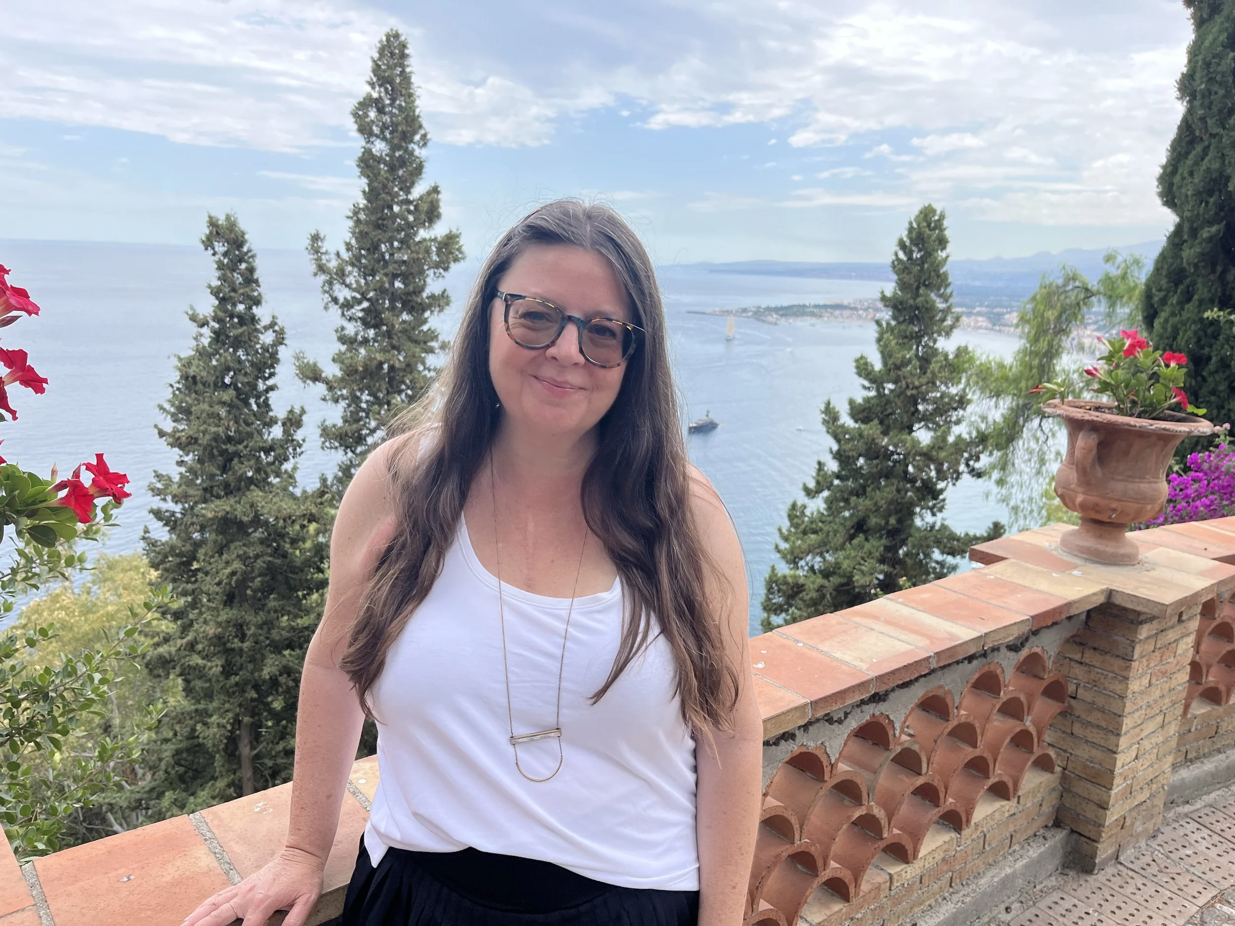 A woman with long brown hair and glasses smiling outdoors with a lake and trees in the background. She is standing by a brick railing with potted flowers.
