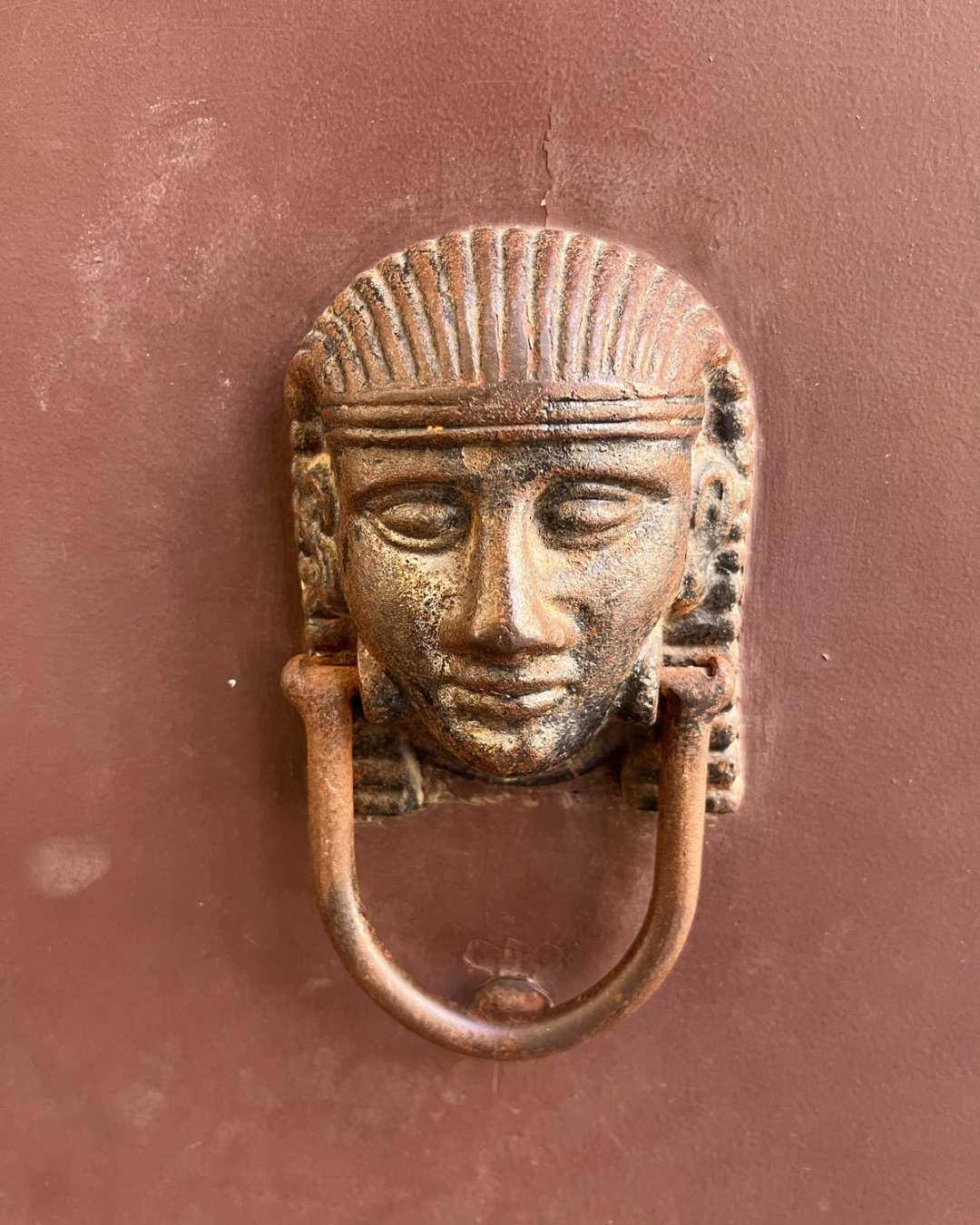 Ancient bronze door knocker in the shape of a human face with traditional jewelry and a headdress, mounted on a pink wall.