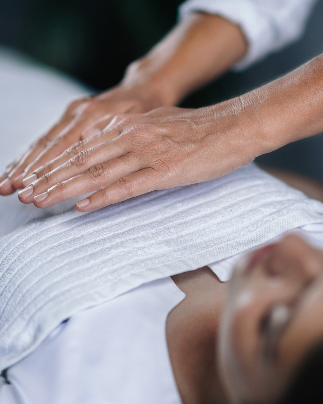Person receiving a massage, lying on a massage table with eyes closed, wearing a white shirt, while a massage therapist applies pressure with hands on the person's chest.