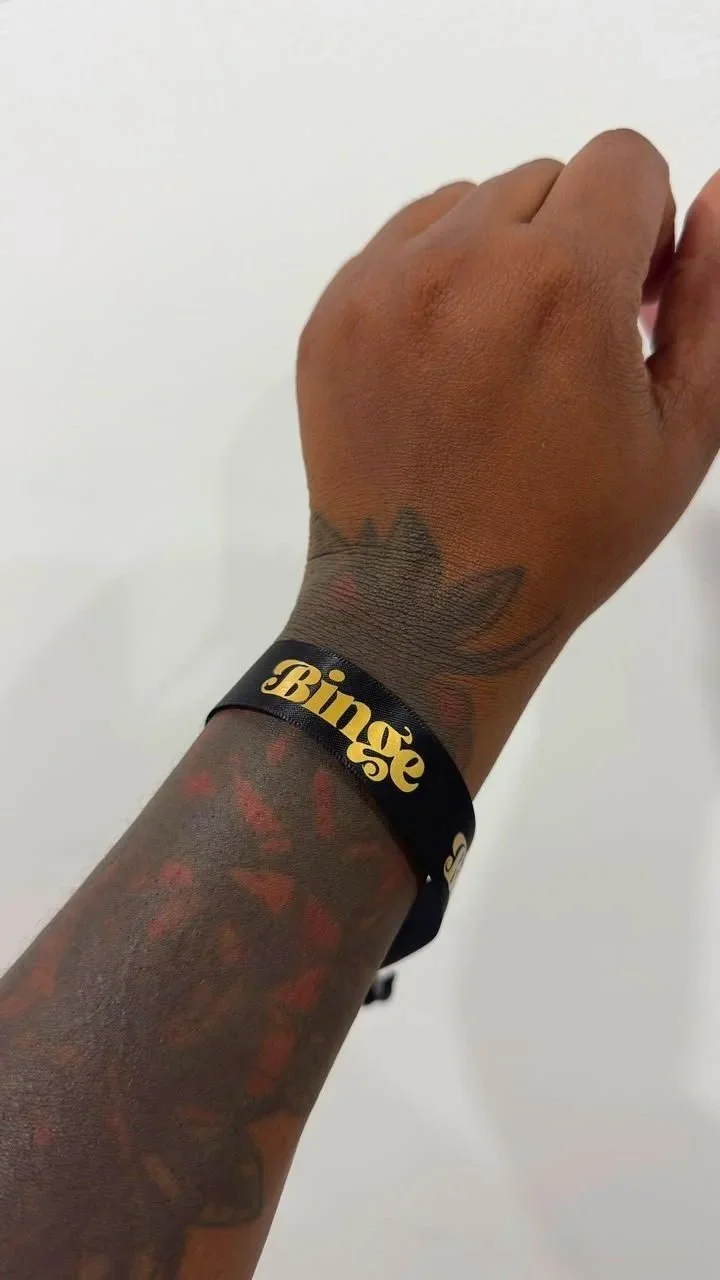 A person's hand and wrist with tattoos, wearing a black wristband that says 'Binge' in gold lettering, against a plain white background.