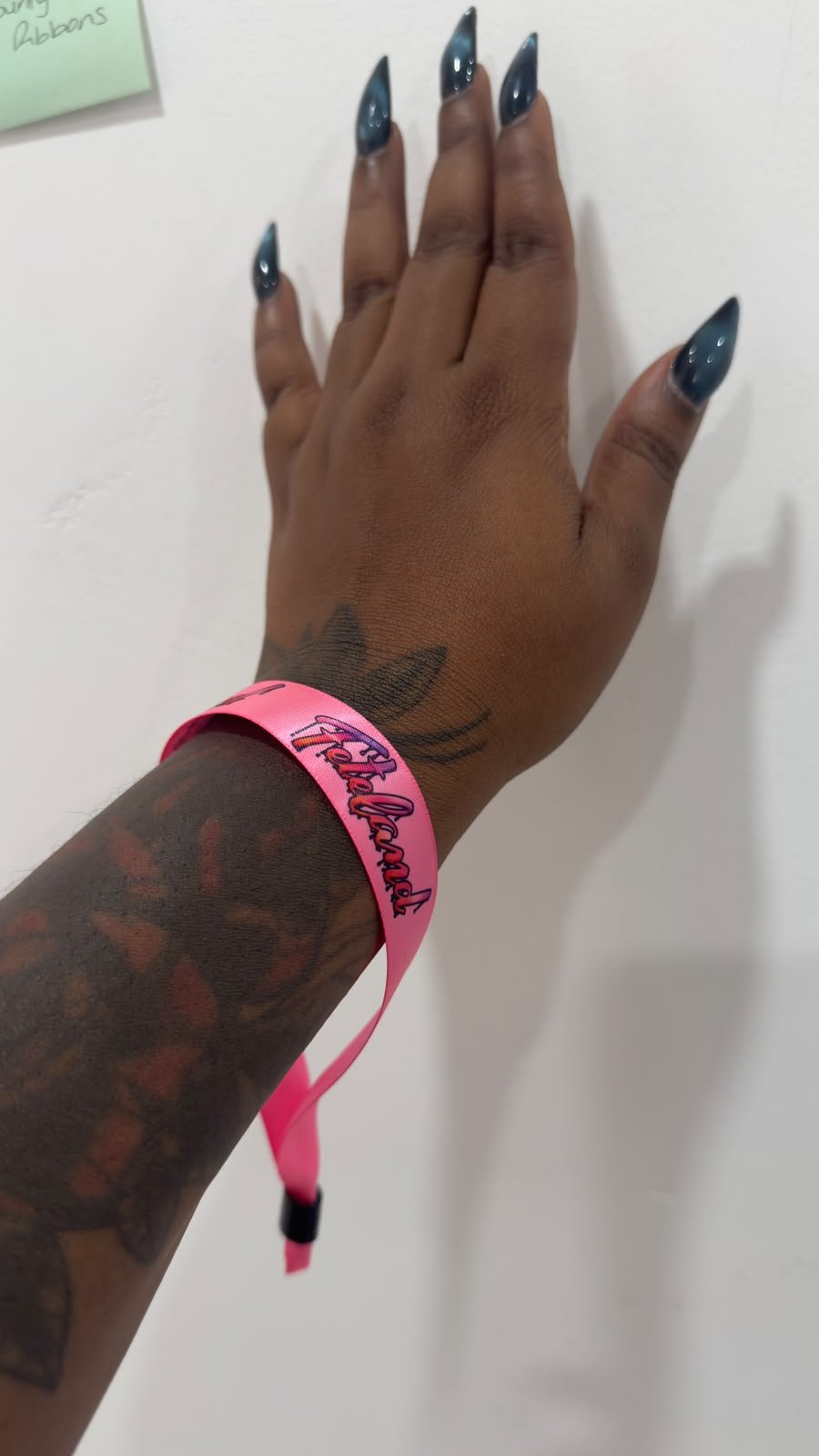 A hand with long, dark blue nails and a tattoo on the wrist, wearing a pink wristband that says 'Feteland' in black and pink lettering, against a white background.