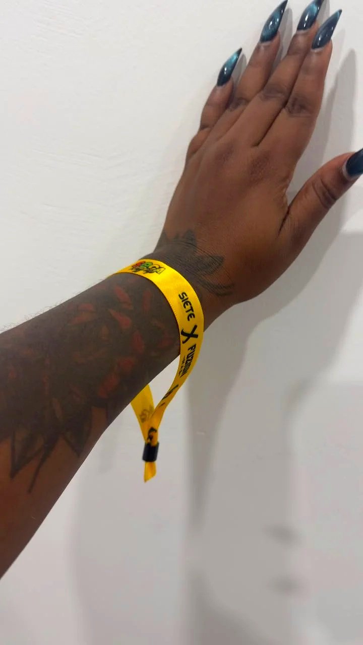 Hand with dark nail polish wearing a yellow event wristband, resting against a white wall.