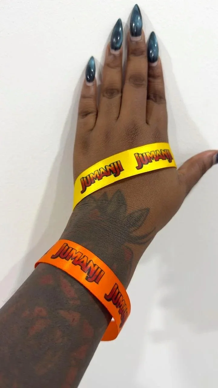 A hand with shiny, dark blue nails and tattoos, wearing yellow and orange wristbands with Jumanji logos, held up against a plain white background.
