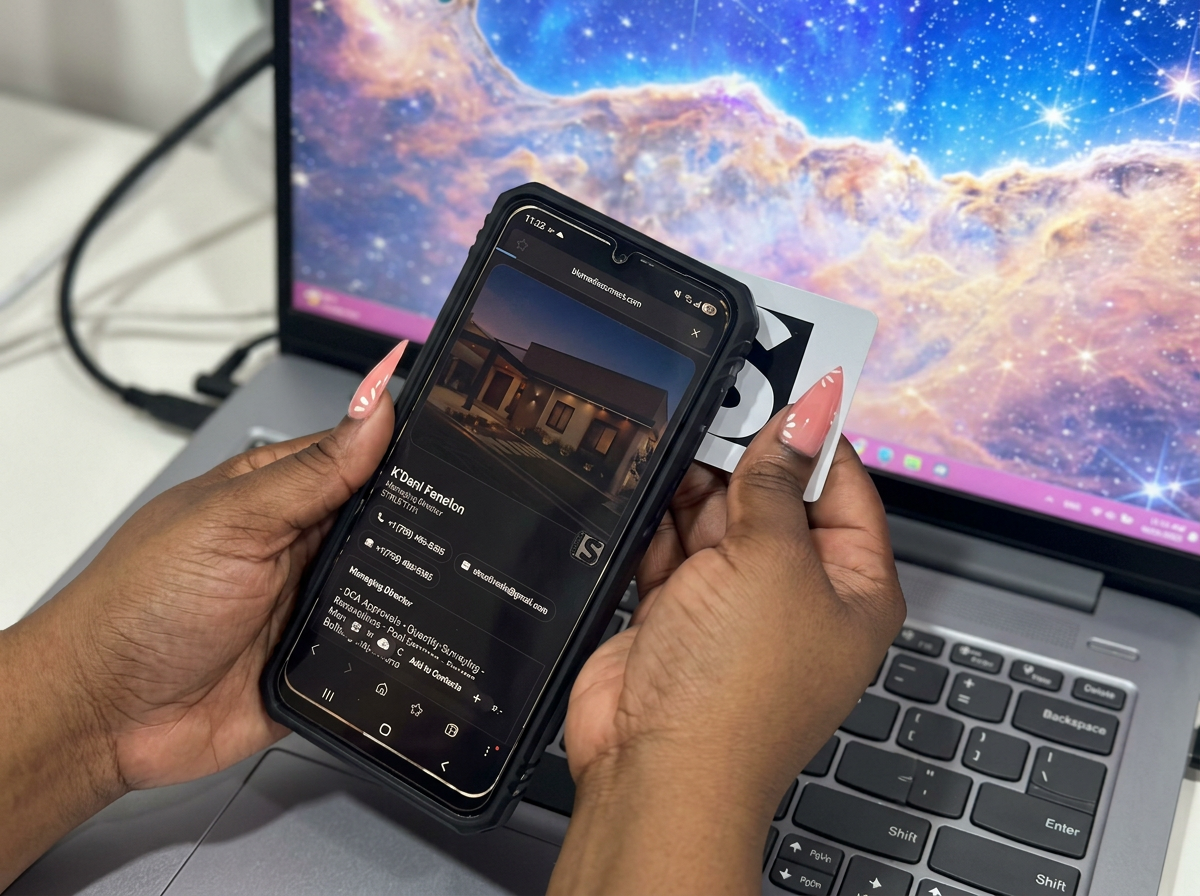 A person with long manicured nails holds a smartphone over a laptop with a galaxy wallpaper on the screen. The phone displays a real estate listing for a house at dusk.