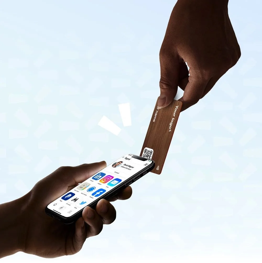 Two hands, one holding a smartphone displaying social media icons, the other holding a wooden NFC card,  scanning or transferring information between them.