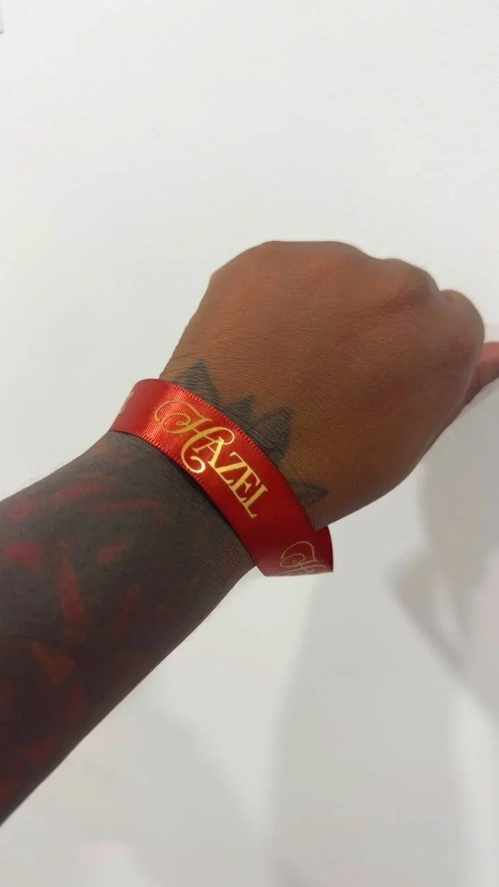 A person's wrist with a red wristband that has gold lettering reading 'Hazel' .