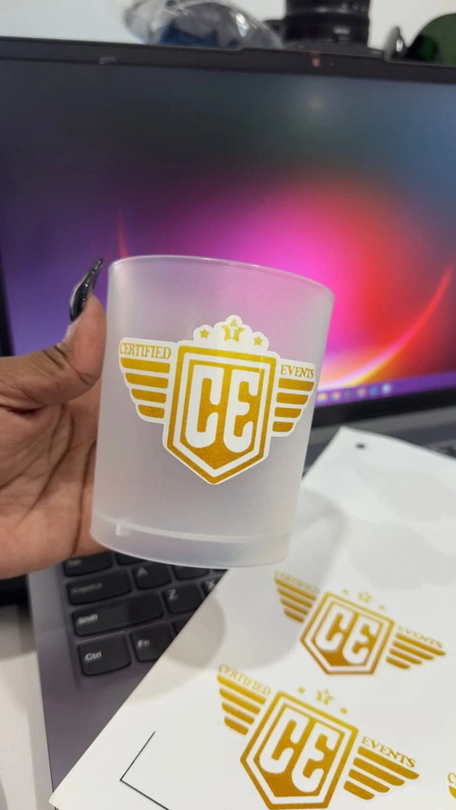 A hand holding a frosted glass with the gold logo of 'Certified Events' and the initials 'CE', with a laptop keyboard, certificate, and computer screen in the background.