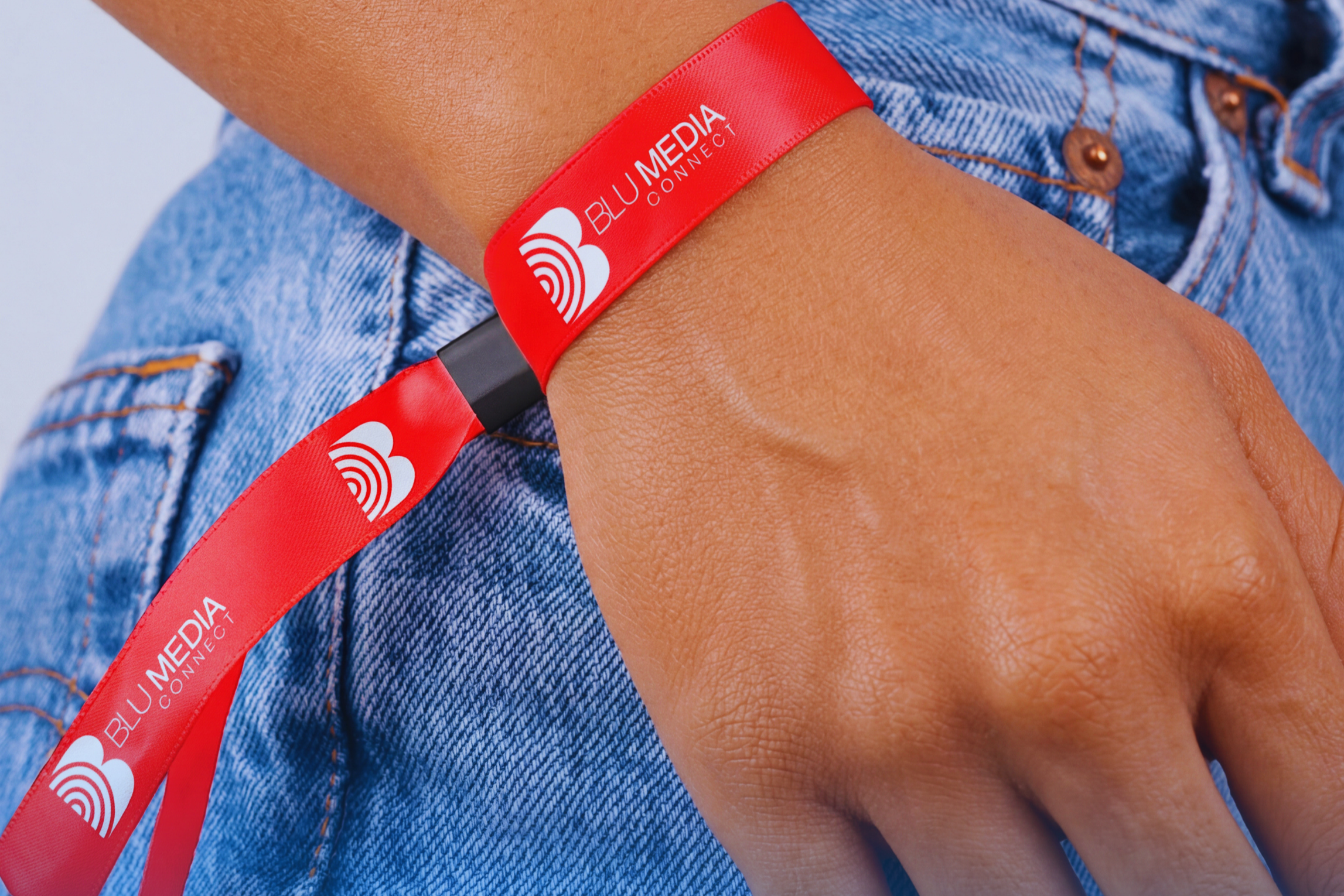 Close-up of a person's arm wearing a red wristband with the logo and text 'BLU MEDIA CONNECT' on it, against a background of blue denim jeans.