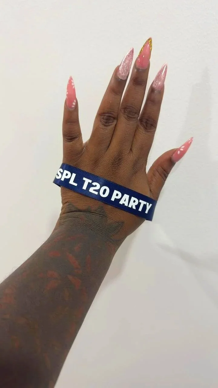 A hand with long, decorated nails and a tattooed arm, wearing a blue wristband that says 'SPL T20 PARTY', is shown against a plain white background.