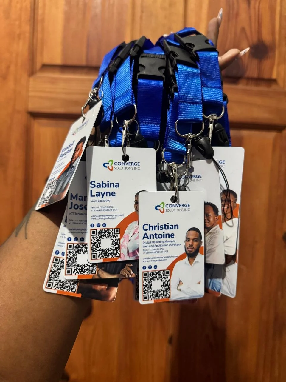 Hand holding numerous blue lanyards with ID badges for Converge Solutions Inc. featuring individual photos, names, job titles, contact information, and QR codes, against a wooden background.