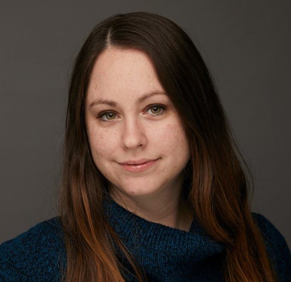 A smiling woman with long brown hair, wearing a dark blue sweater, against a neutral gray background.