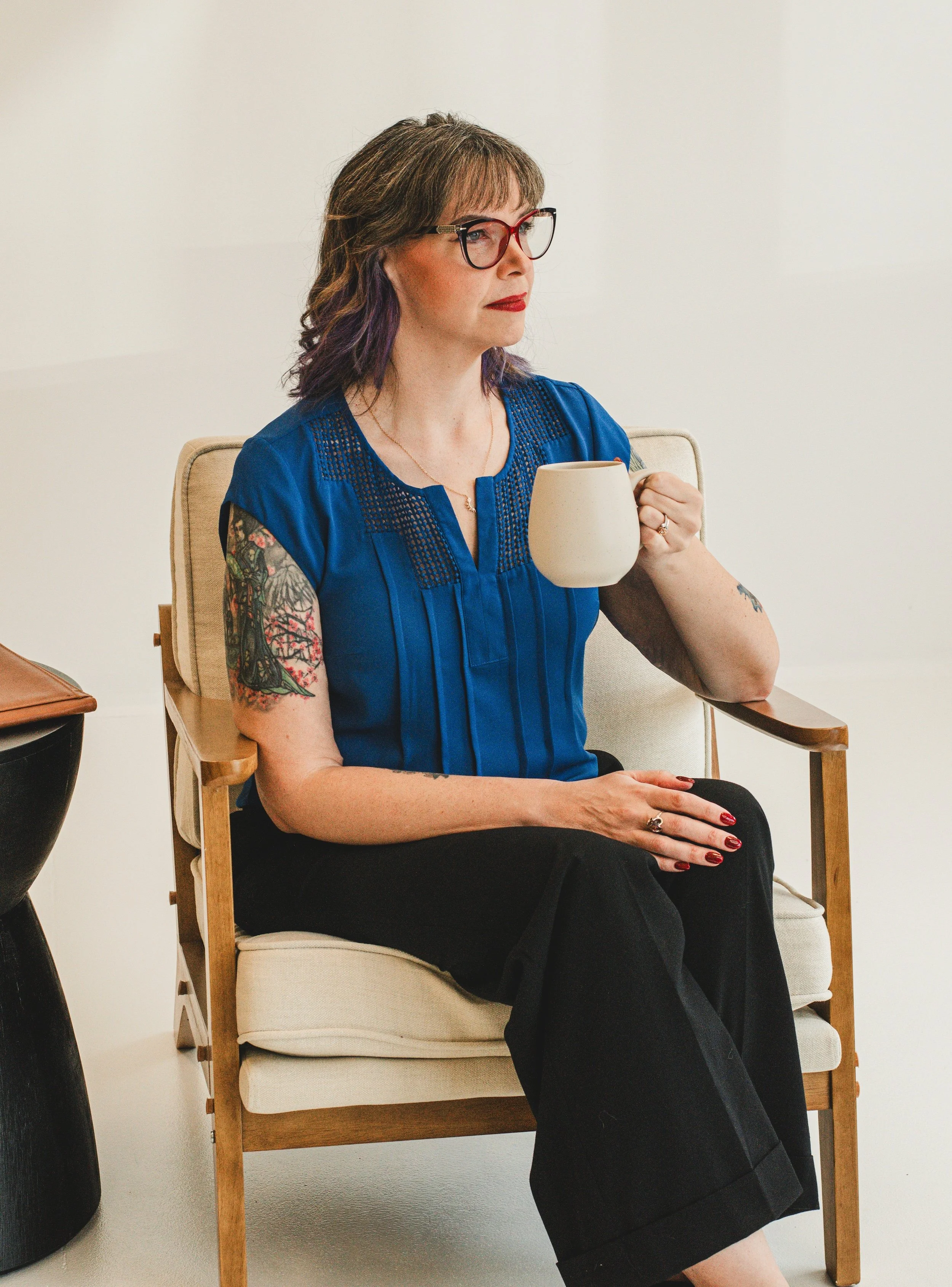 A woman with brown hair and tattoos on her right arm, wearing glasses and a blue top, sitting in a chair holding a white mug.