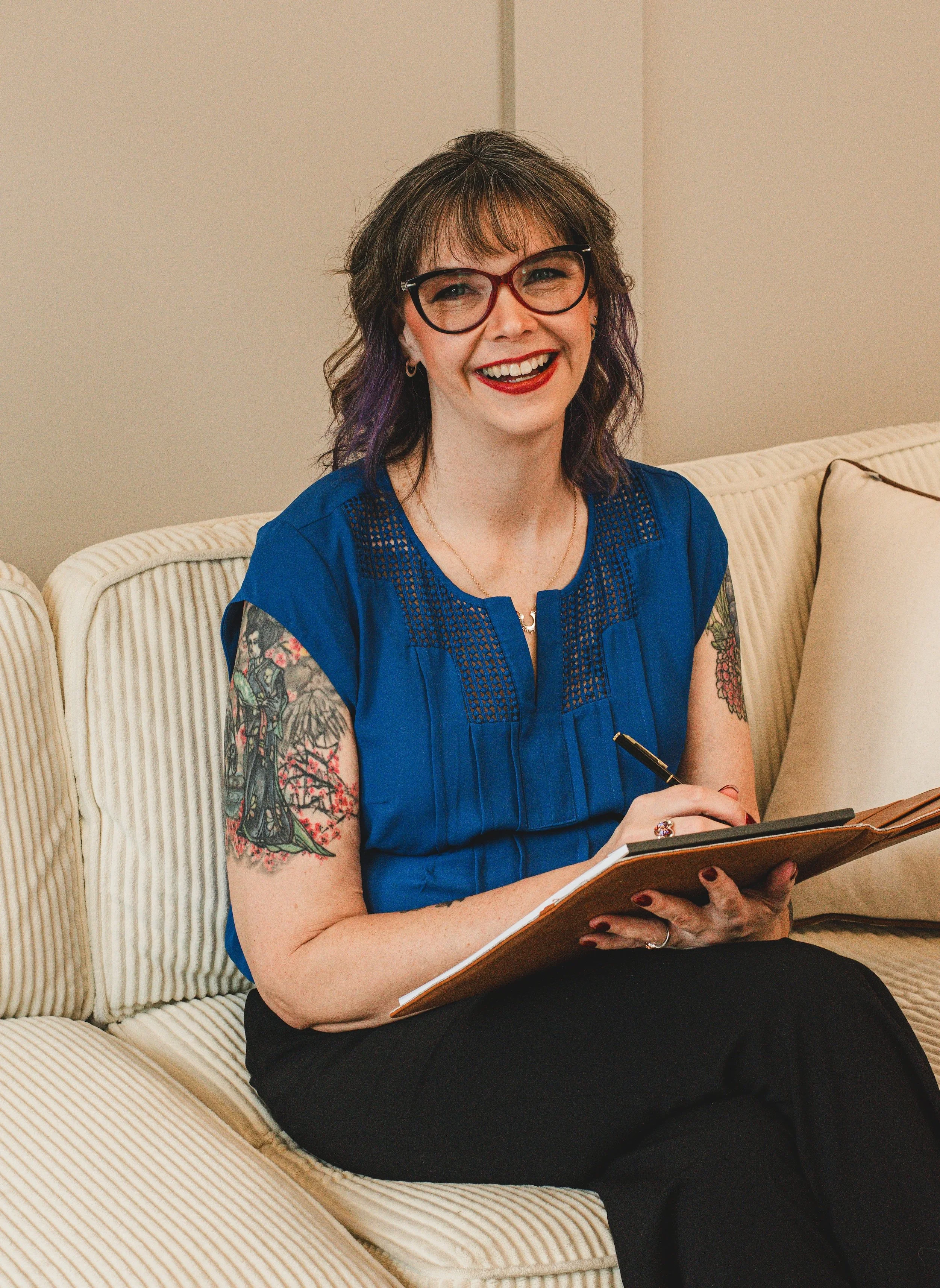A woman with wavy brown hair that has purple tips, wearing glasses and a blue blouse, sitting on a beige sofa, smiling, and holding an open notebook.