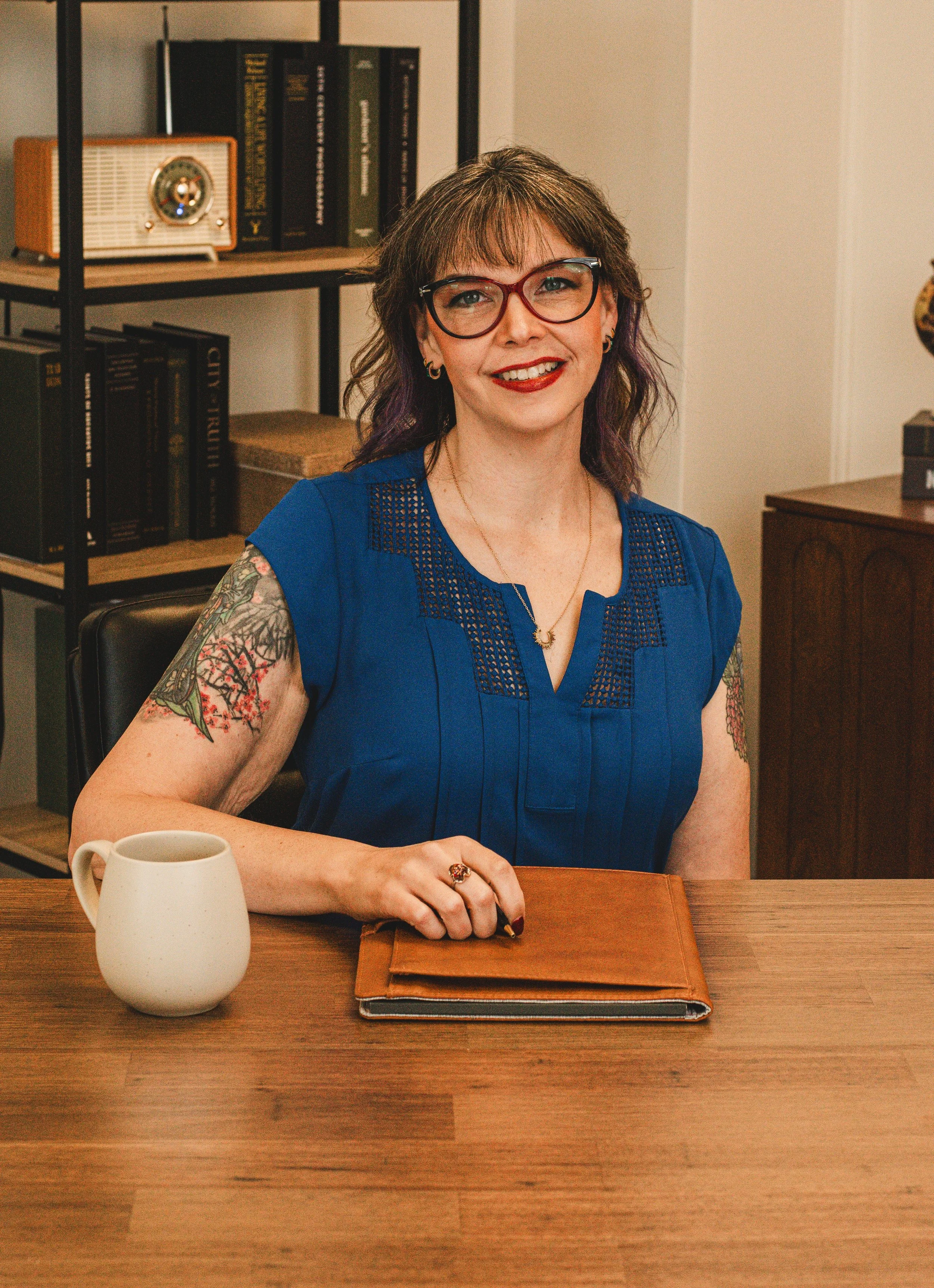 A woman with glasses and tattoos, wearing a blue top, sitting at a wooden table with a white mug and a leather-bound notebook, smiling at the camera, in a room with a bookshelf and a vintage radio in the background.
