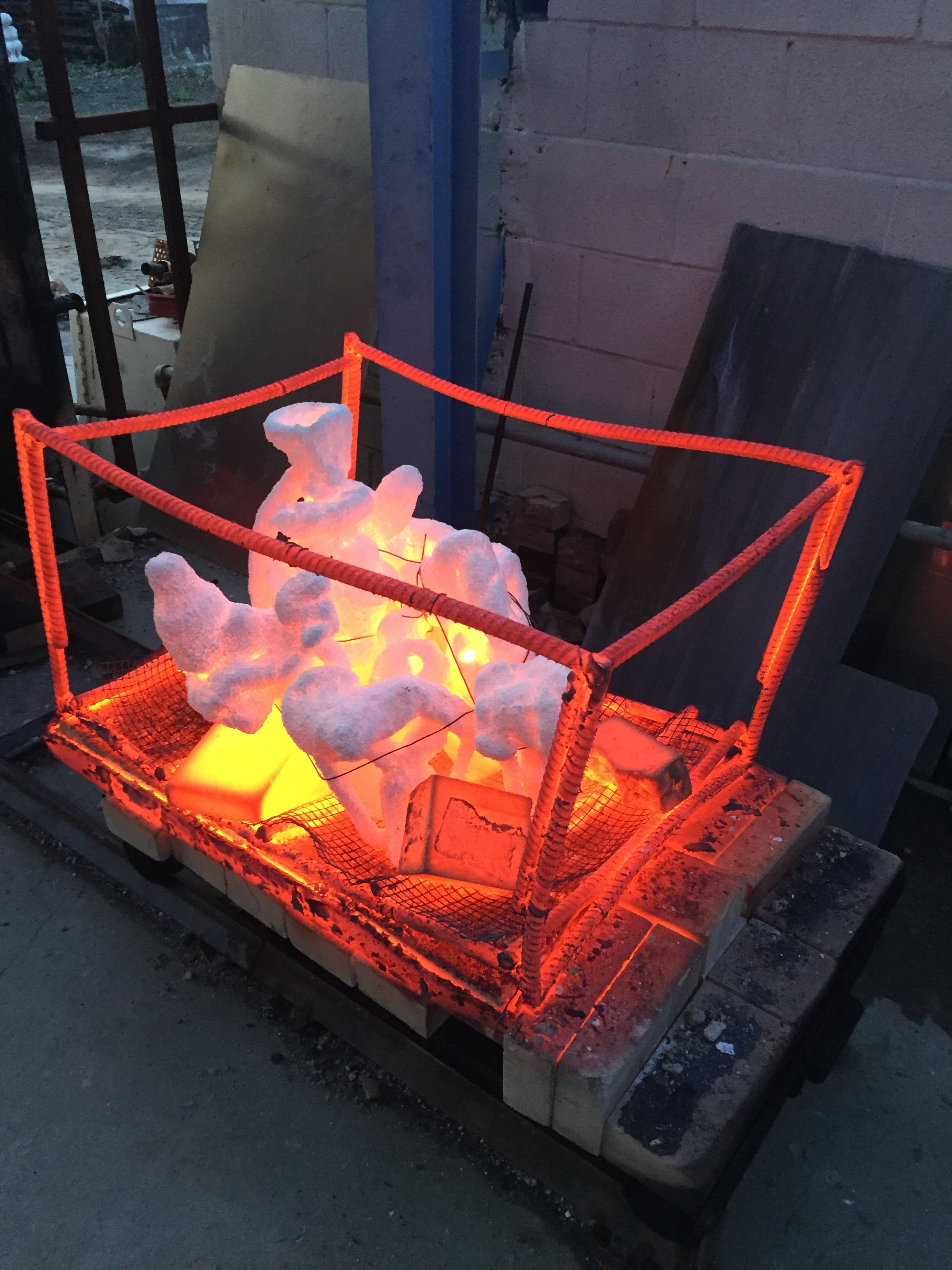 A container of glowing white and orange rocks or minerals, enclosed by a bright orange safety barrier, set on a pallet outside near a brick wall.