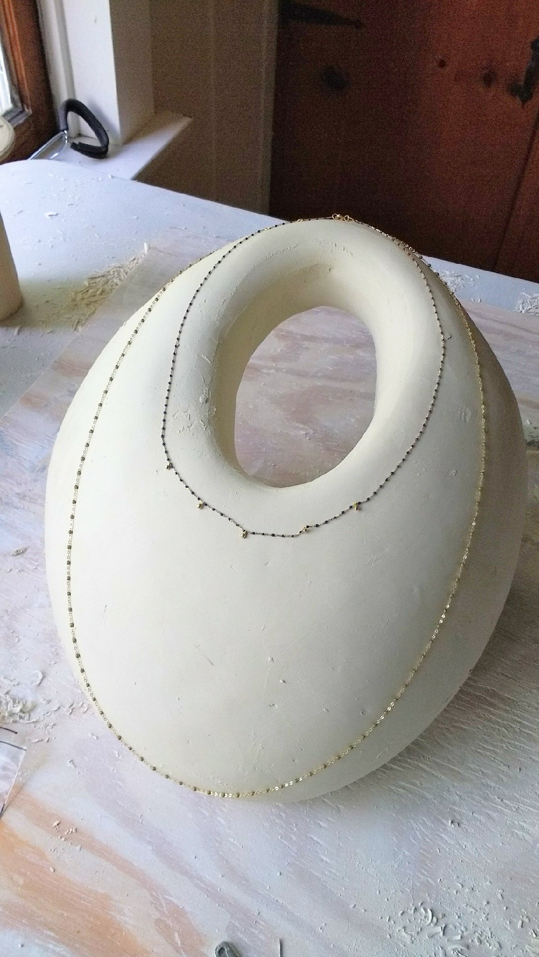 A white, geometric, hollow sculpture with a circular opening in the center, decorated with thin chains draped around it, placed on a dusty wooden surface.