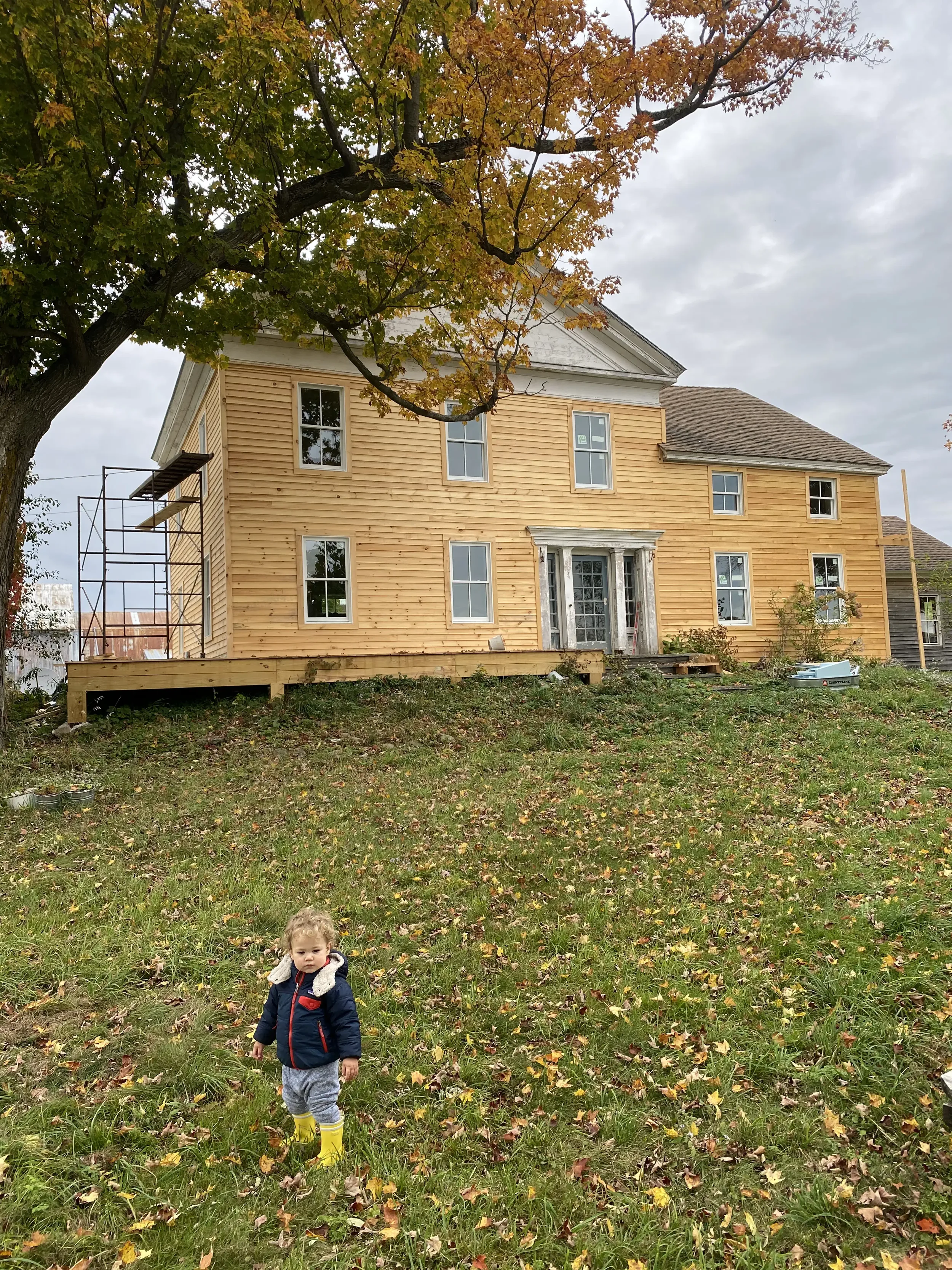 A young child standing on a grassy lawn with fallen leaves, dressed in a navy jacket and yellow rain boots, with a house under construction in the background and a large tree with autumn-colored leaves overhead.