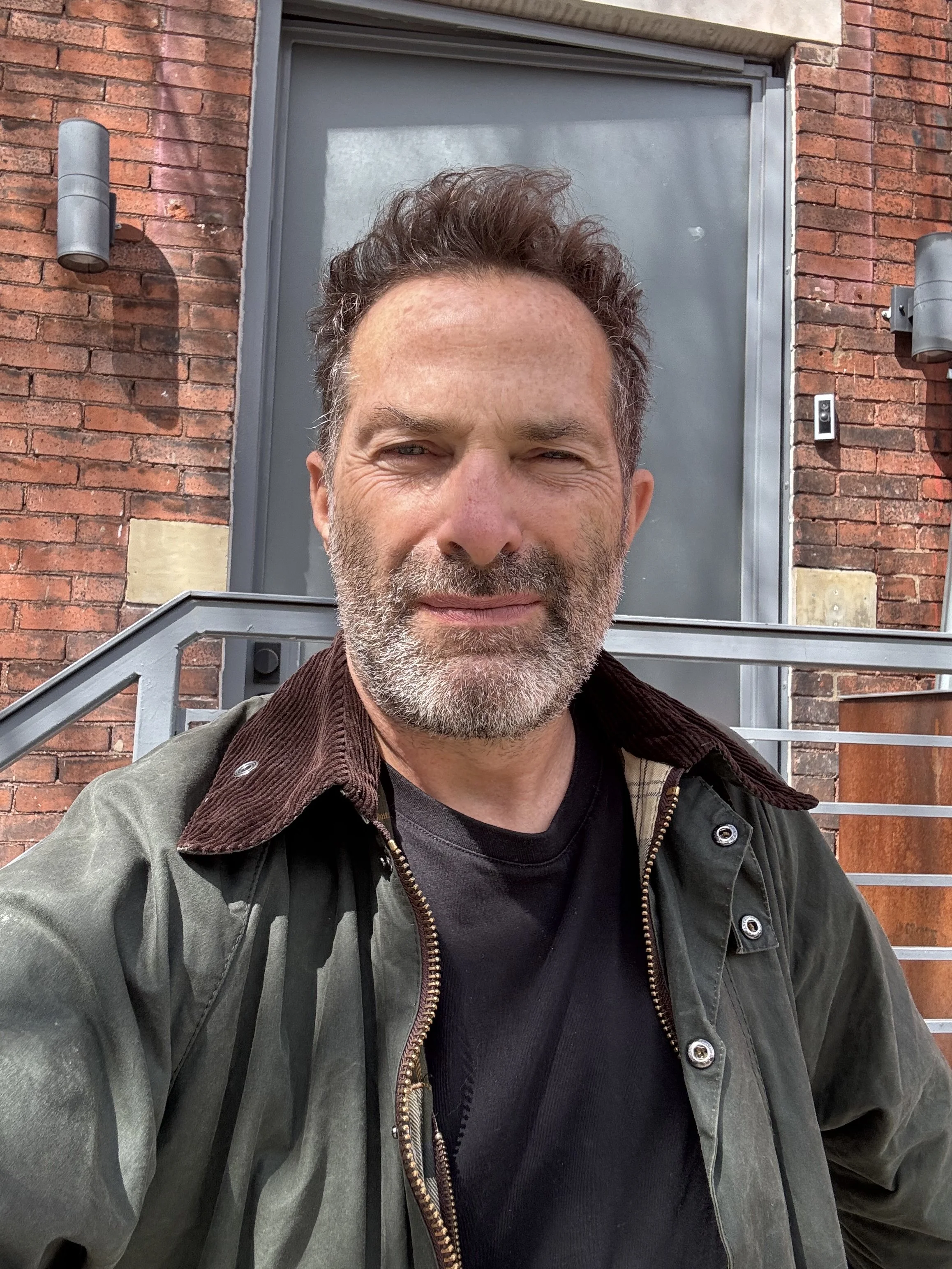 A man with gray facial hair and brown hair, wearing a dark jacket over a black shirt, takes a selfie in front of a gray door set in a brick building. The photo is taken outdoors during the daytime.