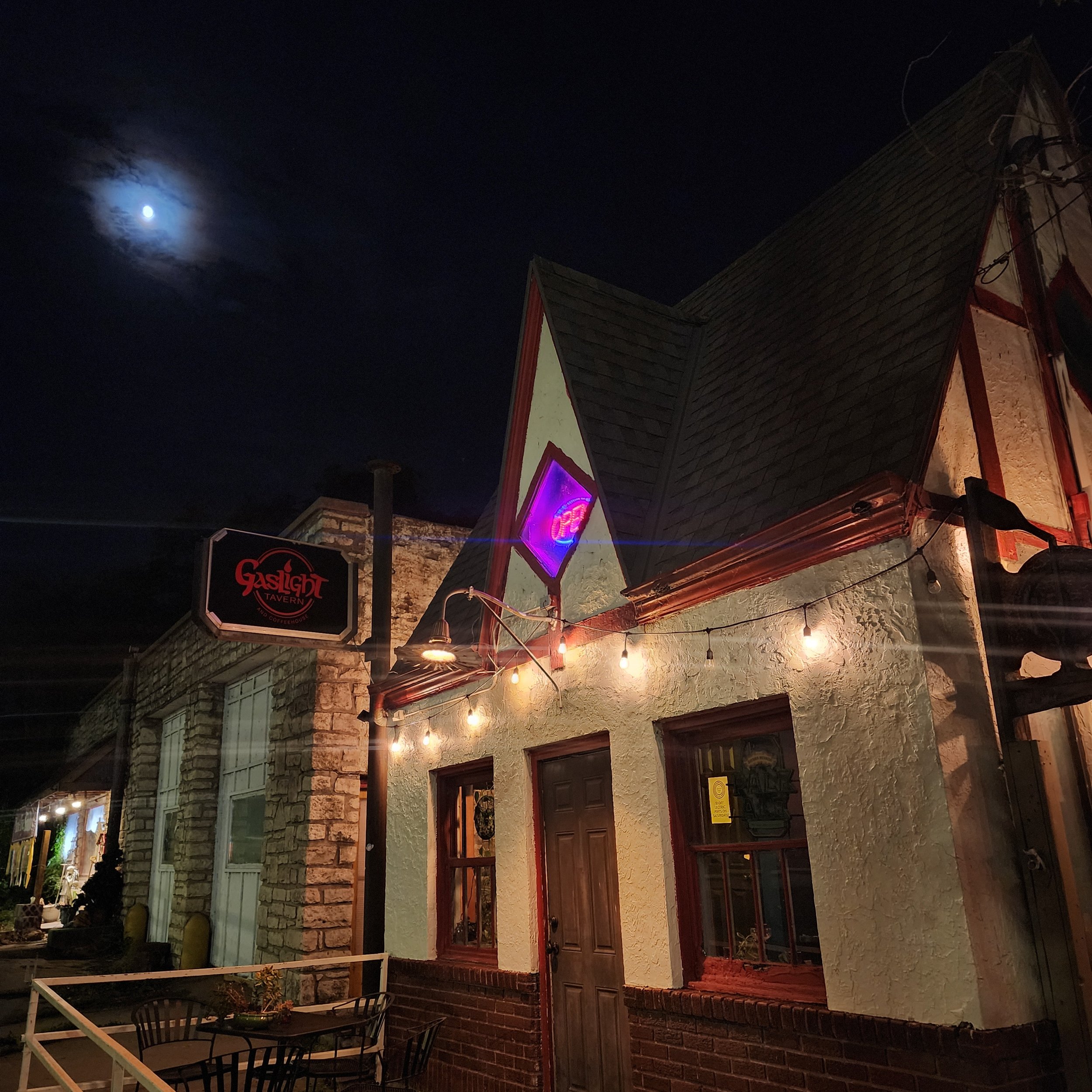 Nighttime scene of a building with a brick and stucco facade, illuminated by string lights, with a neon sign reading 'Gaslight Tavern' and a view of the moon partially obscured by clouds in the dark sky.