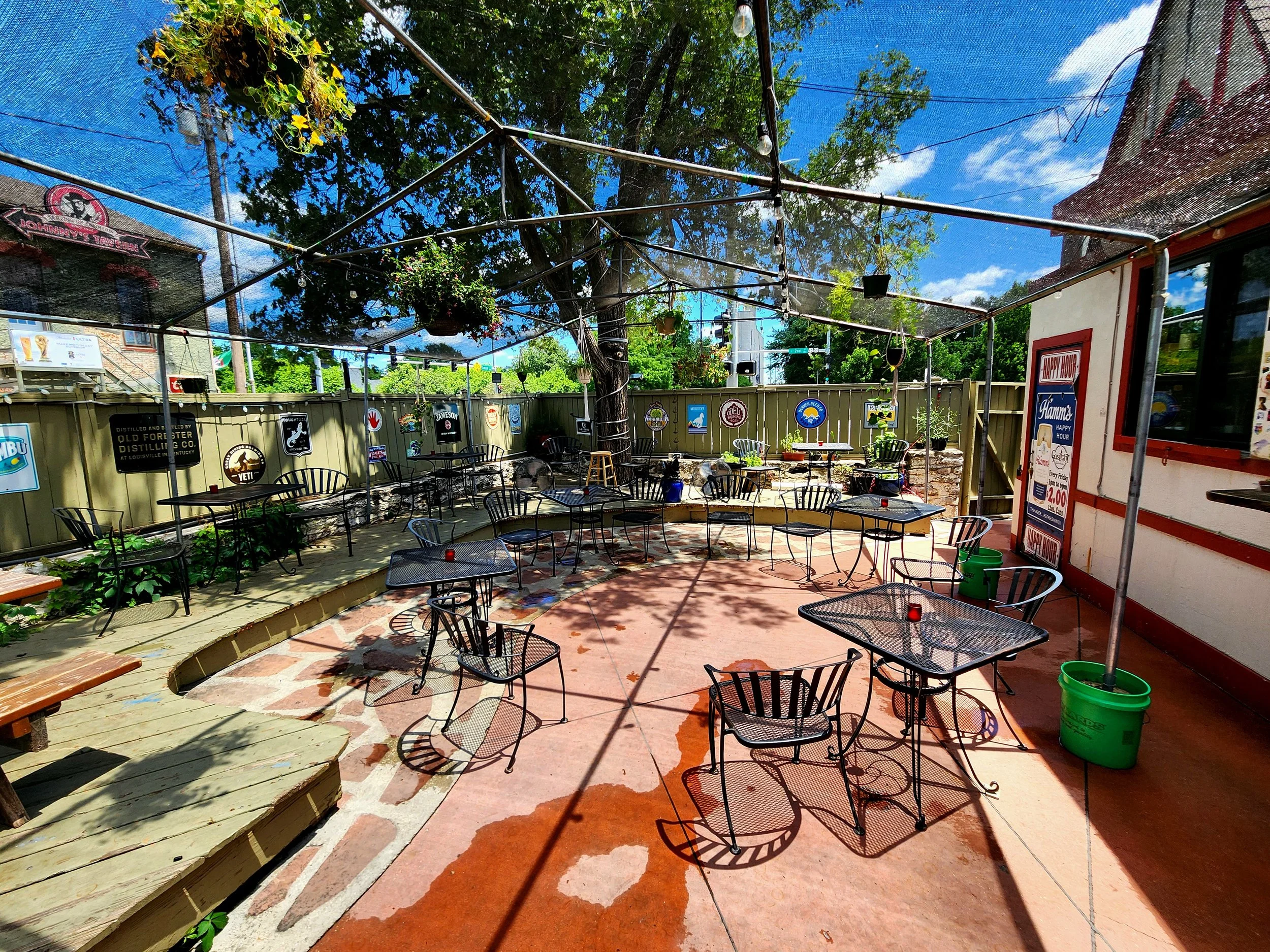 Gaslight Gardens patio area with tables and chairs under a black shade structure decorated with hanging plants and string lights, surrounded by a fence with various signs and posters, on a sunny day with blue skies and green trees.
