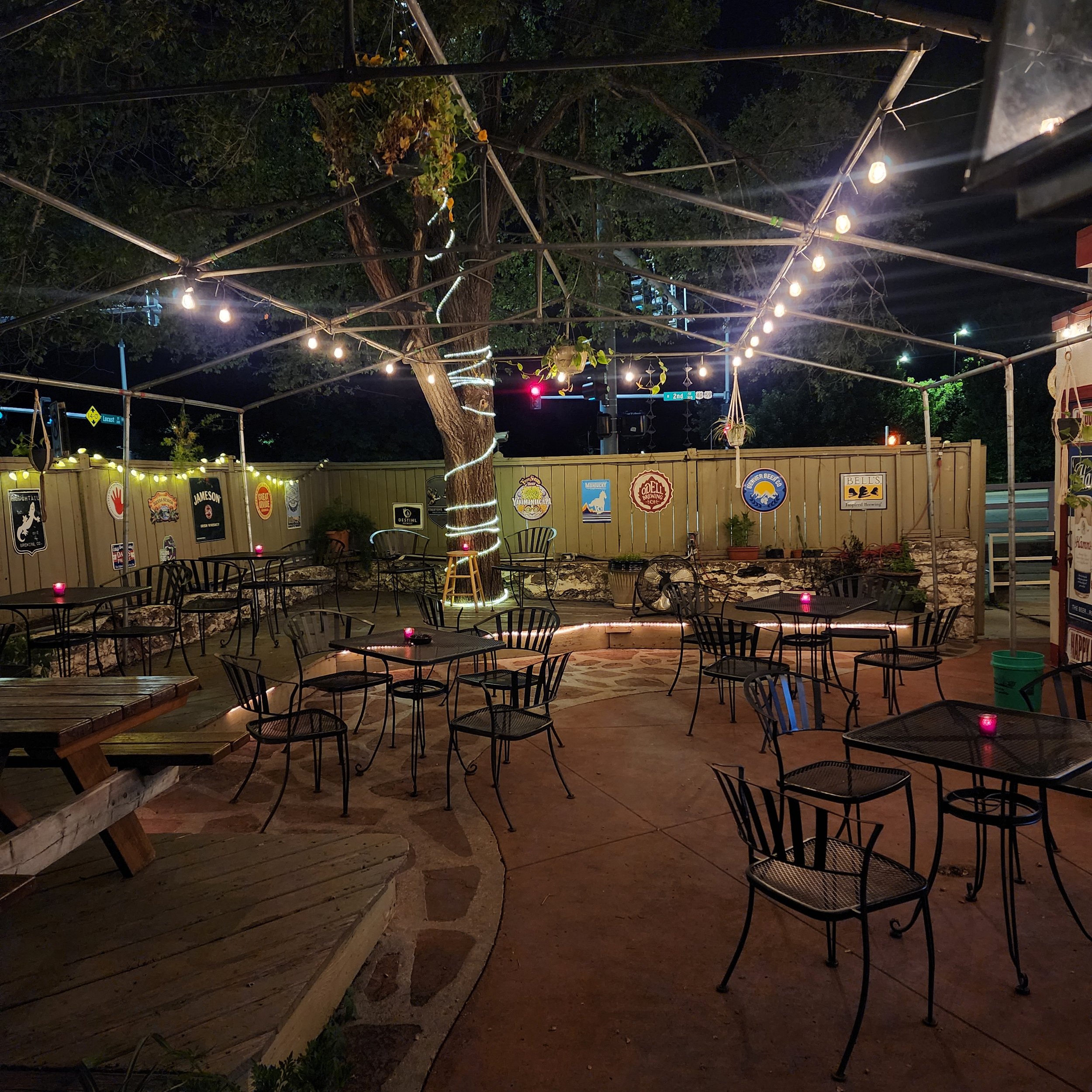 Gaslight Gardens patio area at night with string lights, metal chairs, and tables, surrounded by a wooden fence with signs, and a large tree in the background.