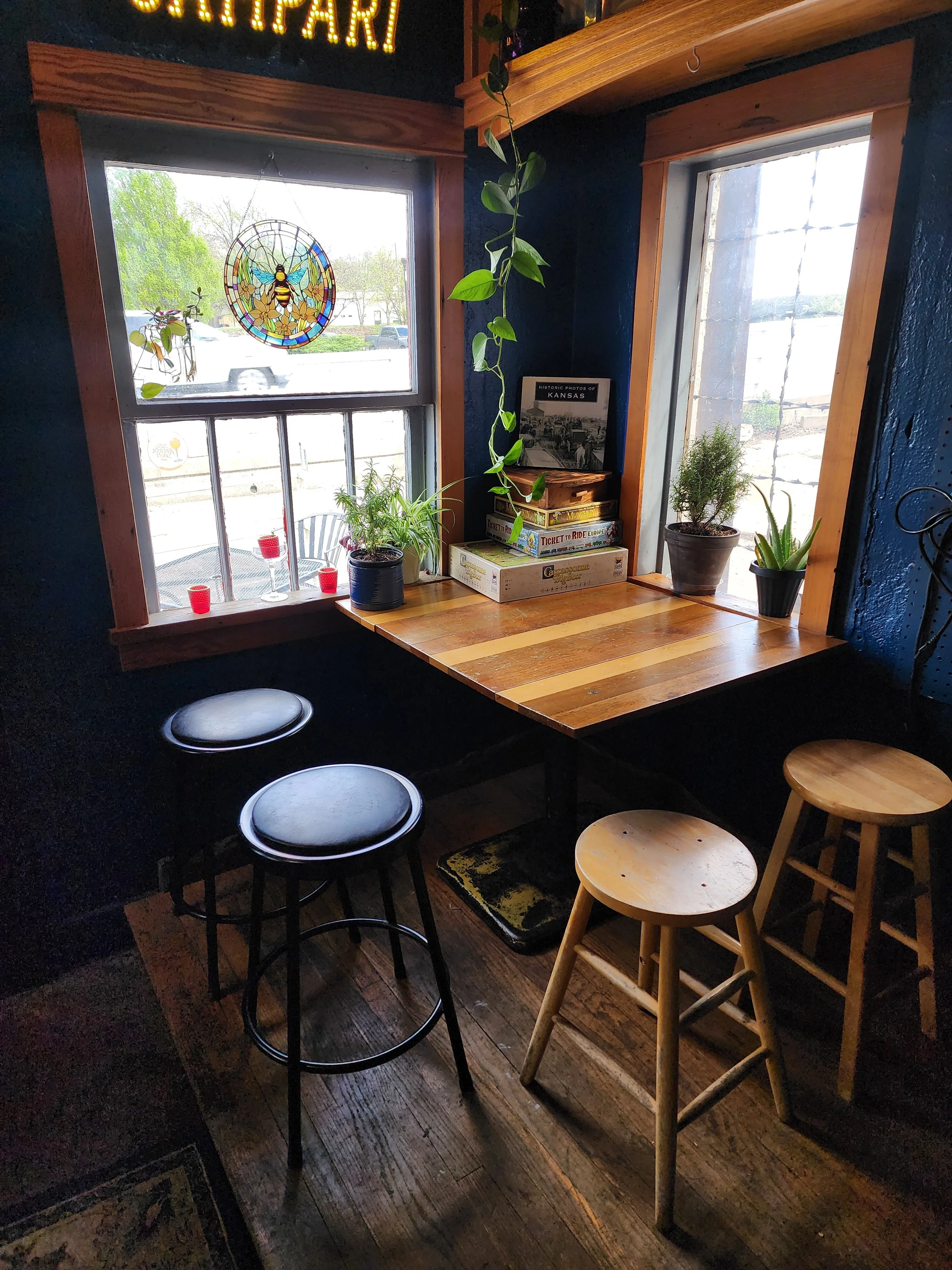 A view of a cozy corner in a cafe with a wooden counter, three stools, and potted plants near the window. There are board games stacked on the counter and a stained-glass bee design in the window. Sunlight streams in from outside, illuminating the sp