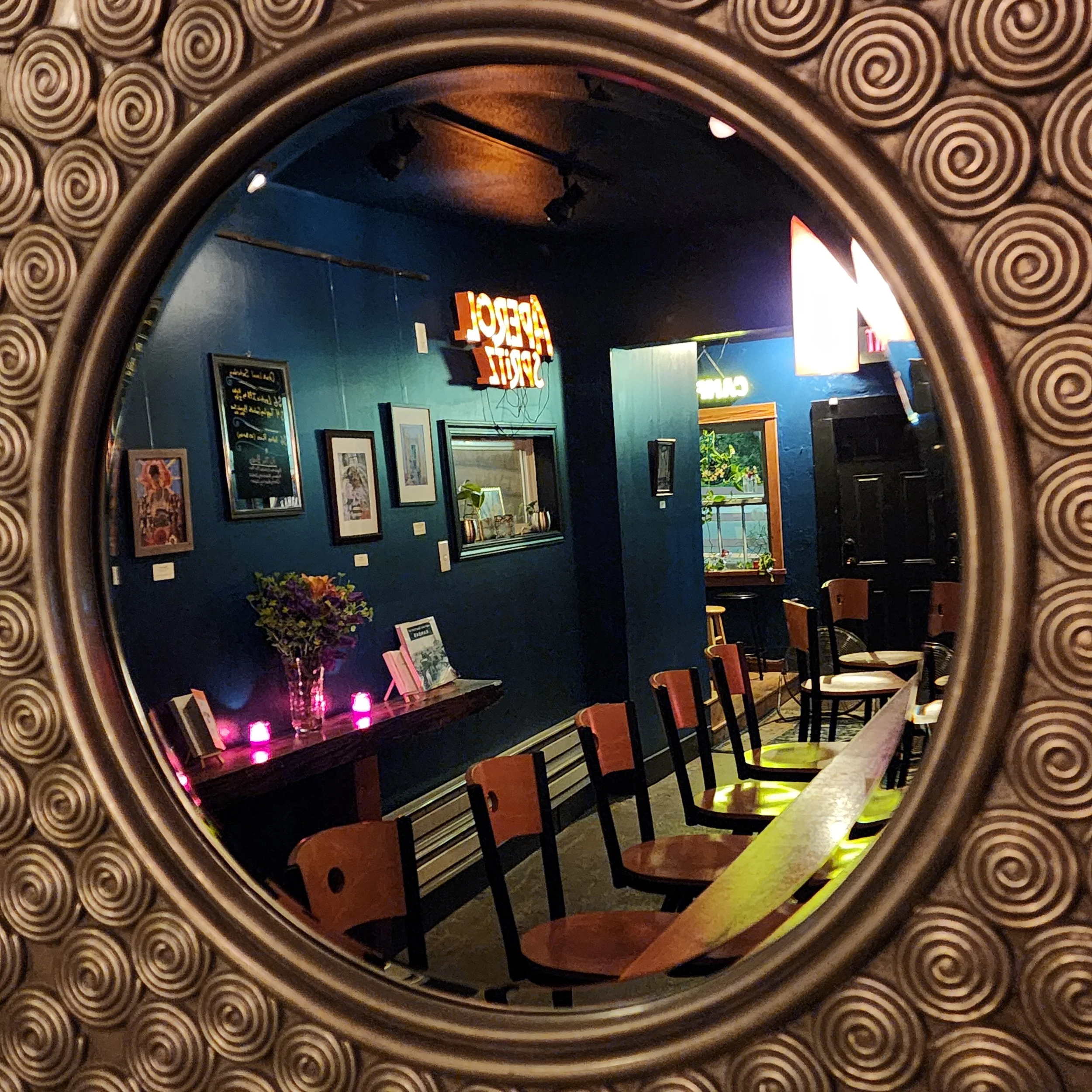 A mirror with a decorative spiral pattern frame reflecting a room with dark blue walls, framed pictures, a table with flowers, a menu, and a neon sign inside a restaurant or cafe.