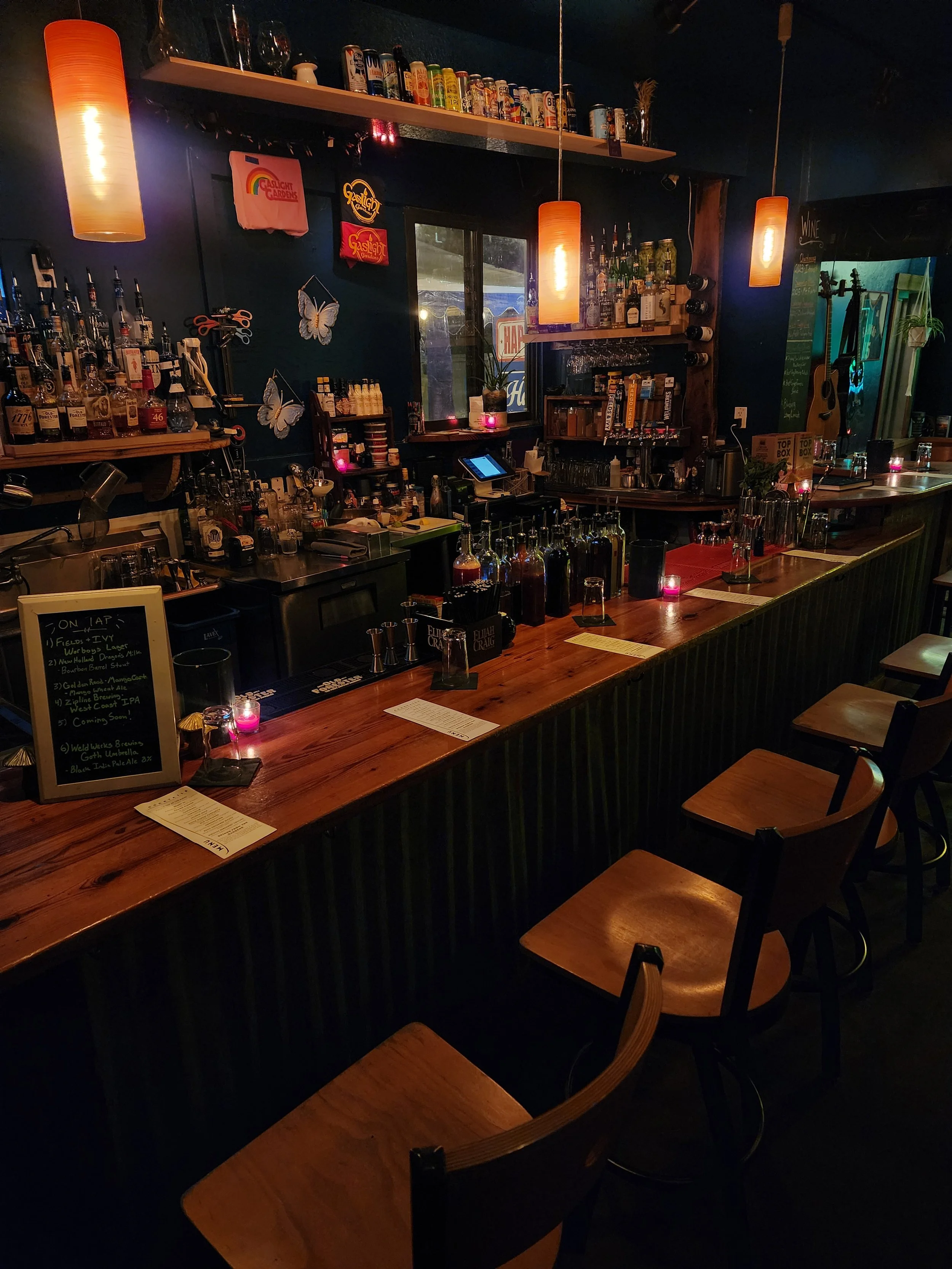 A cozy bar with a wooden bar counter, various bottles of alcohol, glasses, and a menu on the counter. Decorative butterfly wall art, hanging lights, a window, and a chalkboard menu are visible in a dimly lit setting.
