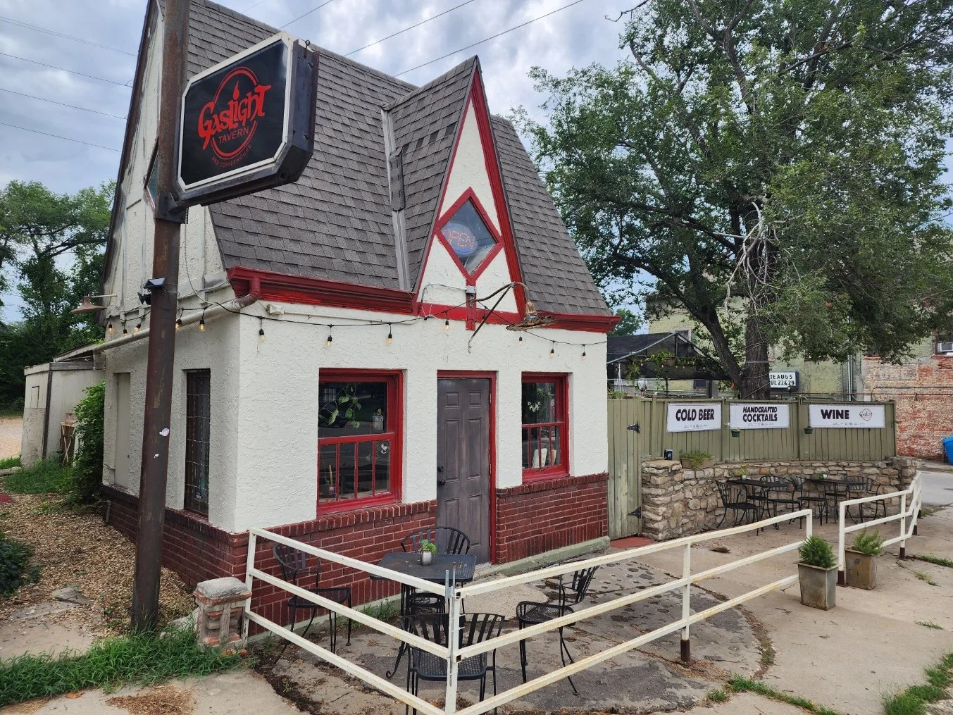 Small, quaint building with white and red trim, used as a tavern or bar, with an outdoor seating area and string lights, and signs indicating drinks such as cold beer, wine, and handcrafted cocktails, in a neighborhood setting.