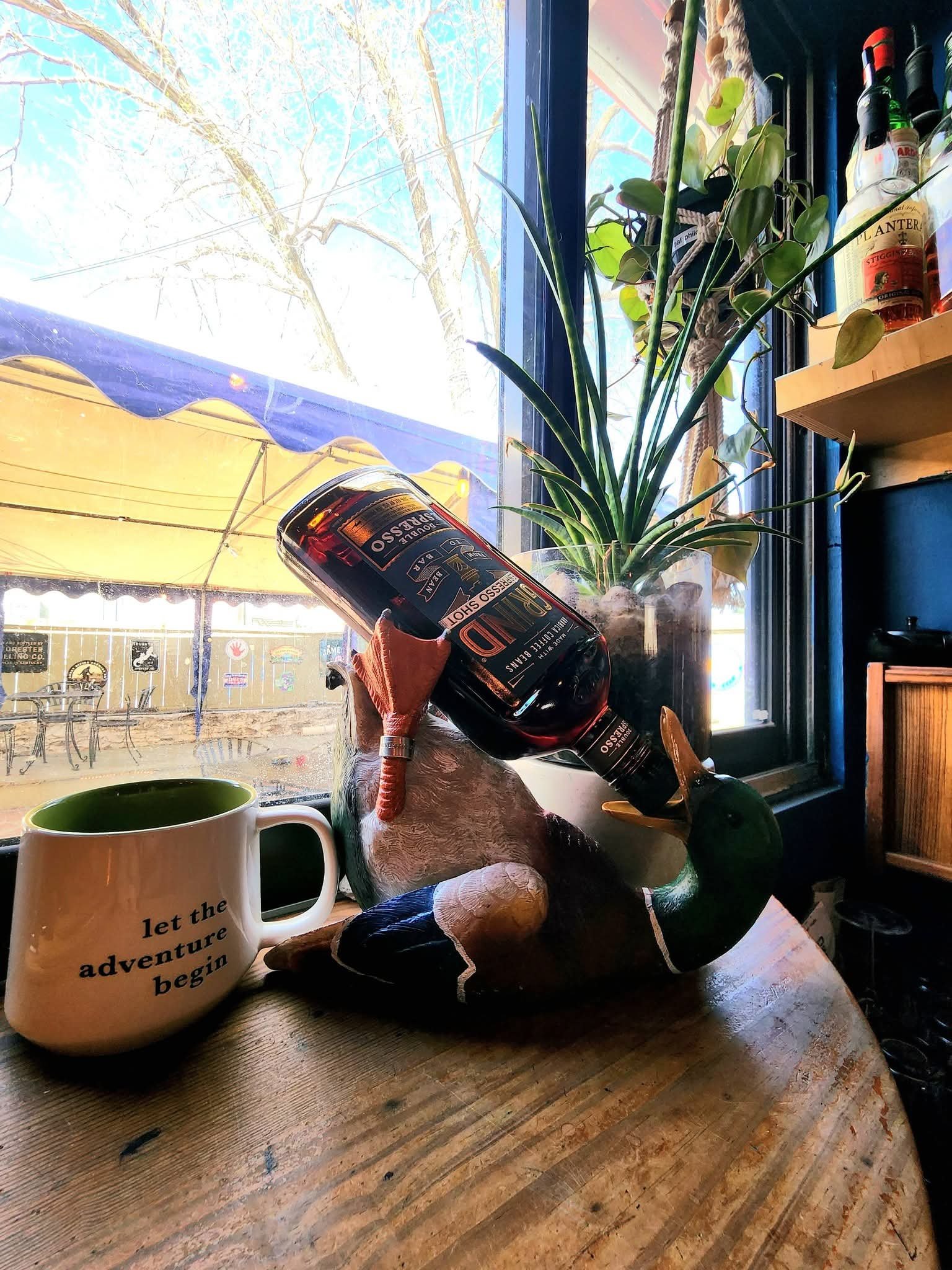 A ceramic mug with the quote "Let the adventure begin" in black text on a white background, placed on a wooden table beside a decorative fox figurine wearing a duck hat, in front of a window with a view of outdoor patio furniture and a yellow awning,