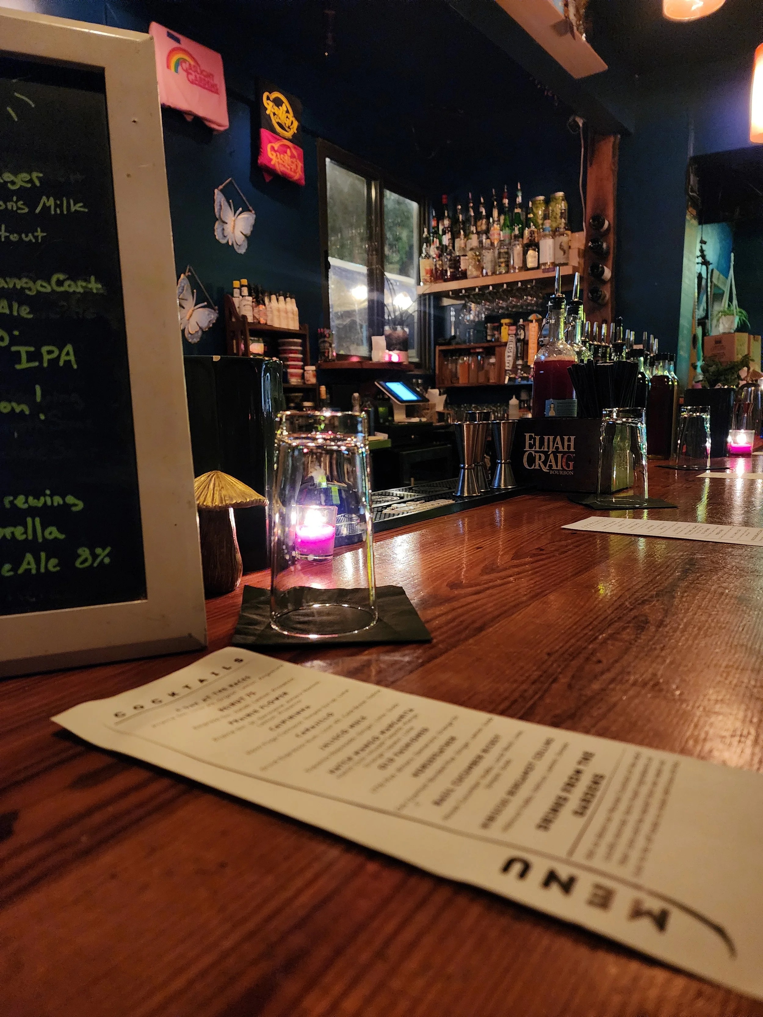 A bar with various bottles of alcohol, a menu, and a blackboard with drink specials in a dimly lit setting with candles.