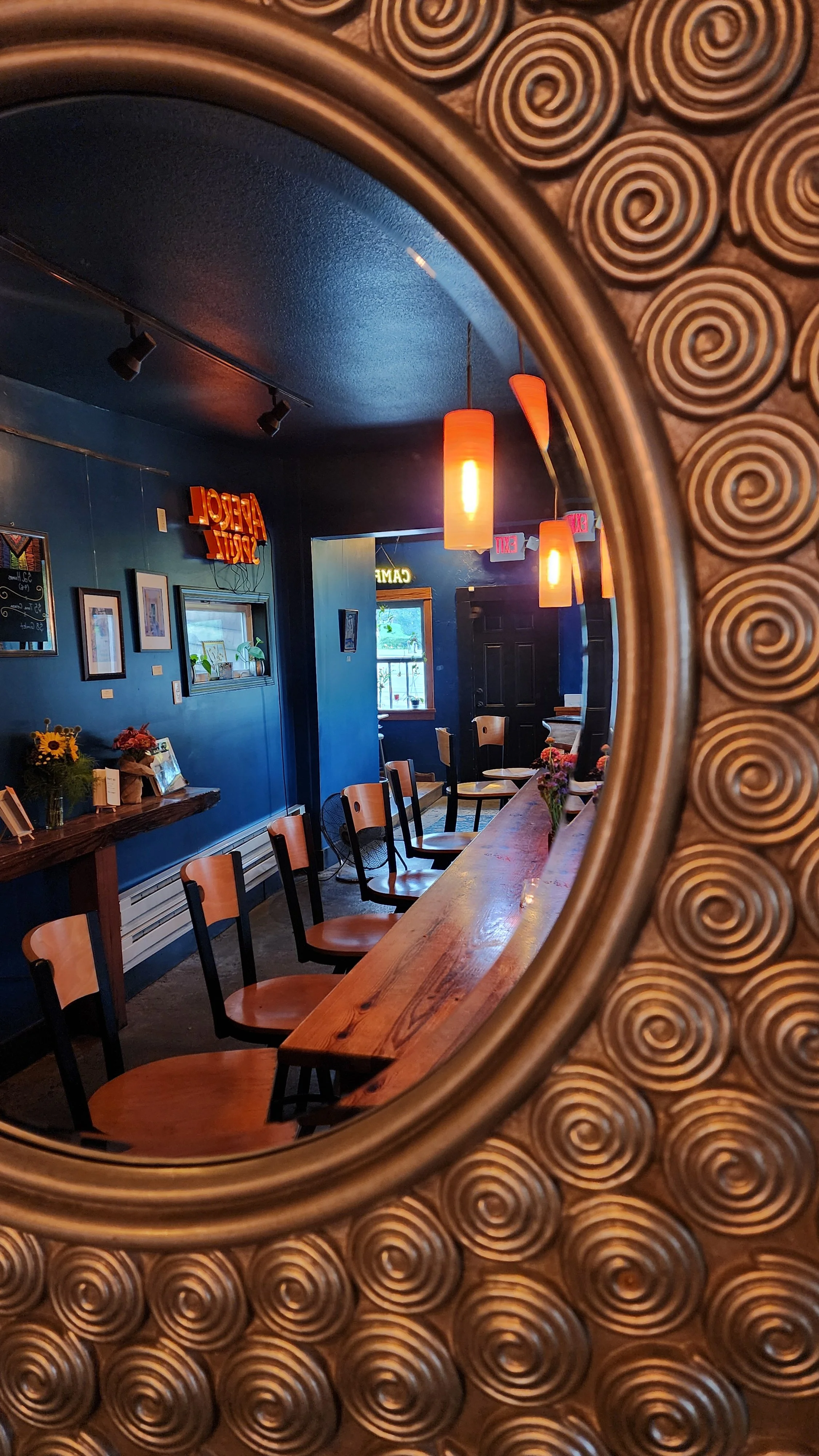 View of a cozy restaurant interior through a decorative round mirror with a swirl pattern. The restaurant has wooden chairs, a wooden bar or counter, pendant lighting, blue walls, framed pictures, and a window with plants.