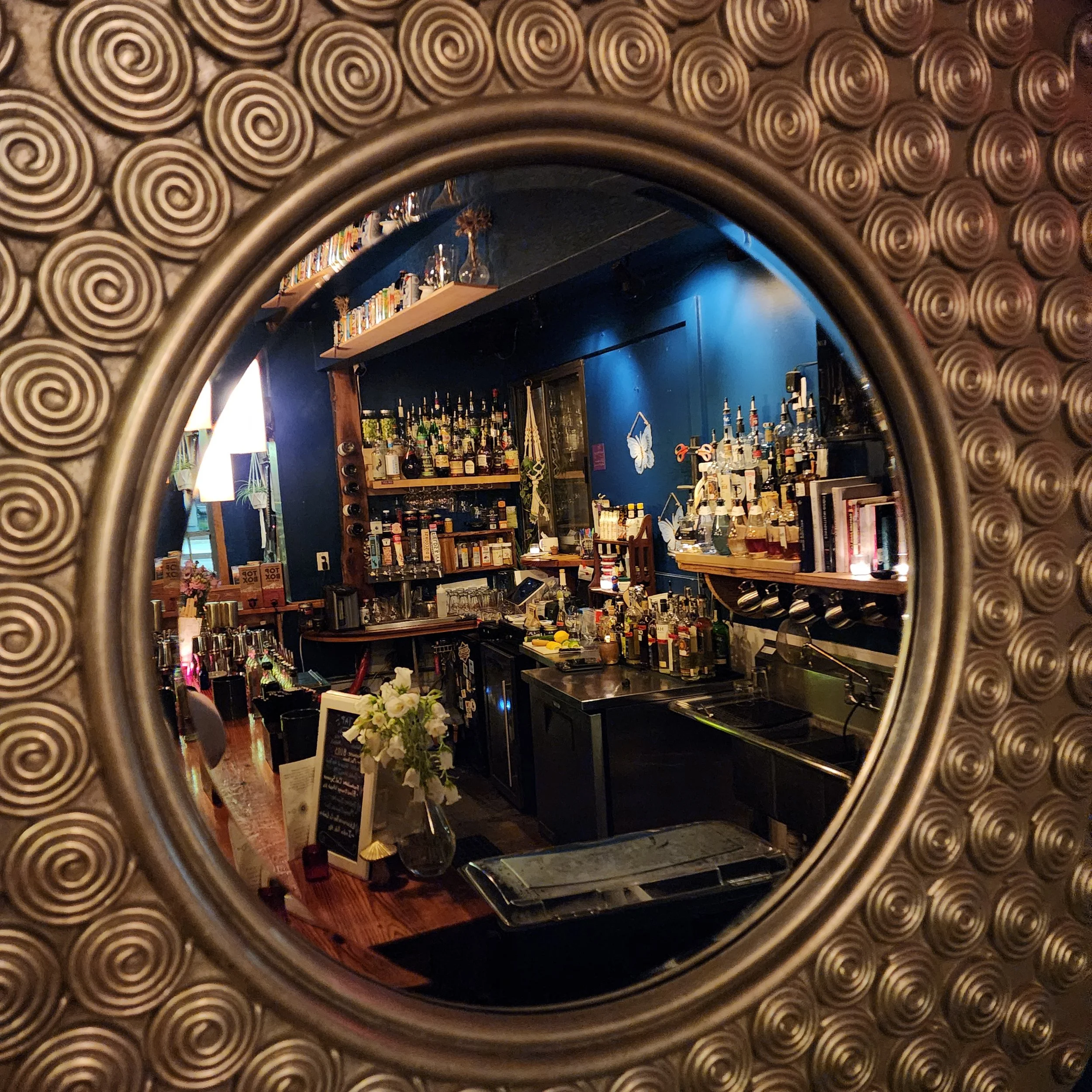 The image shows a bar area with shelves filled with bottles of alcohol, drinks, and bar equipment. The photo is taken through a decorative mirror with an ornate spiral border.