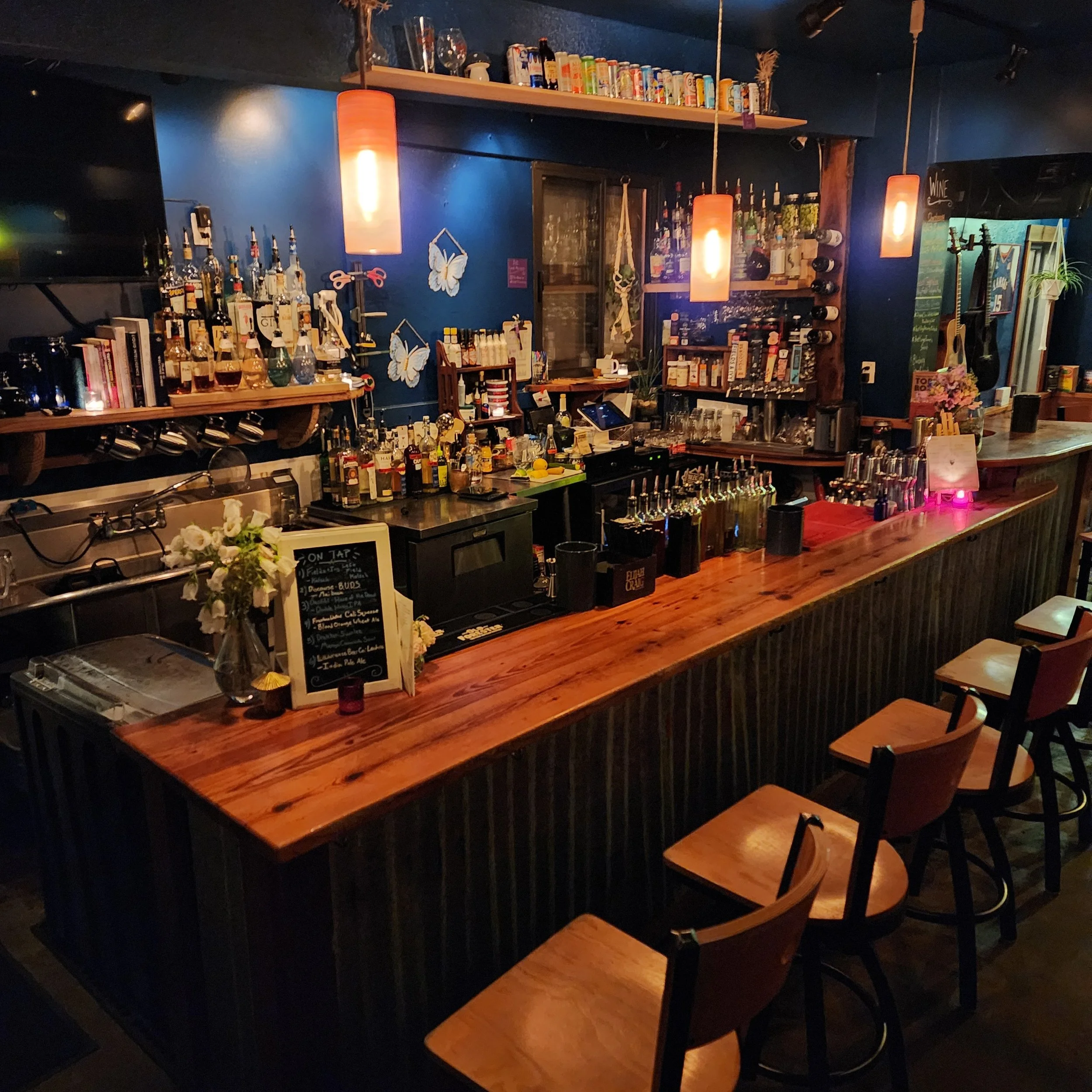 A wooden bar counter with chairs, bottles, and barware in a dimly lit establishment with blue walls and hanging orange lights.