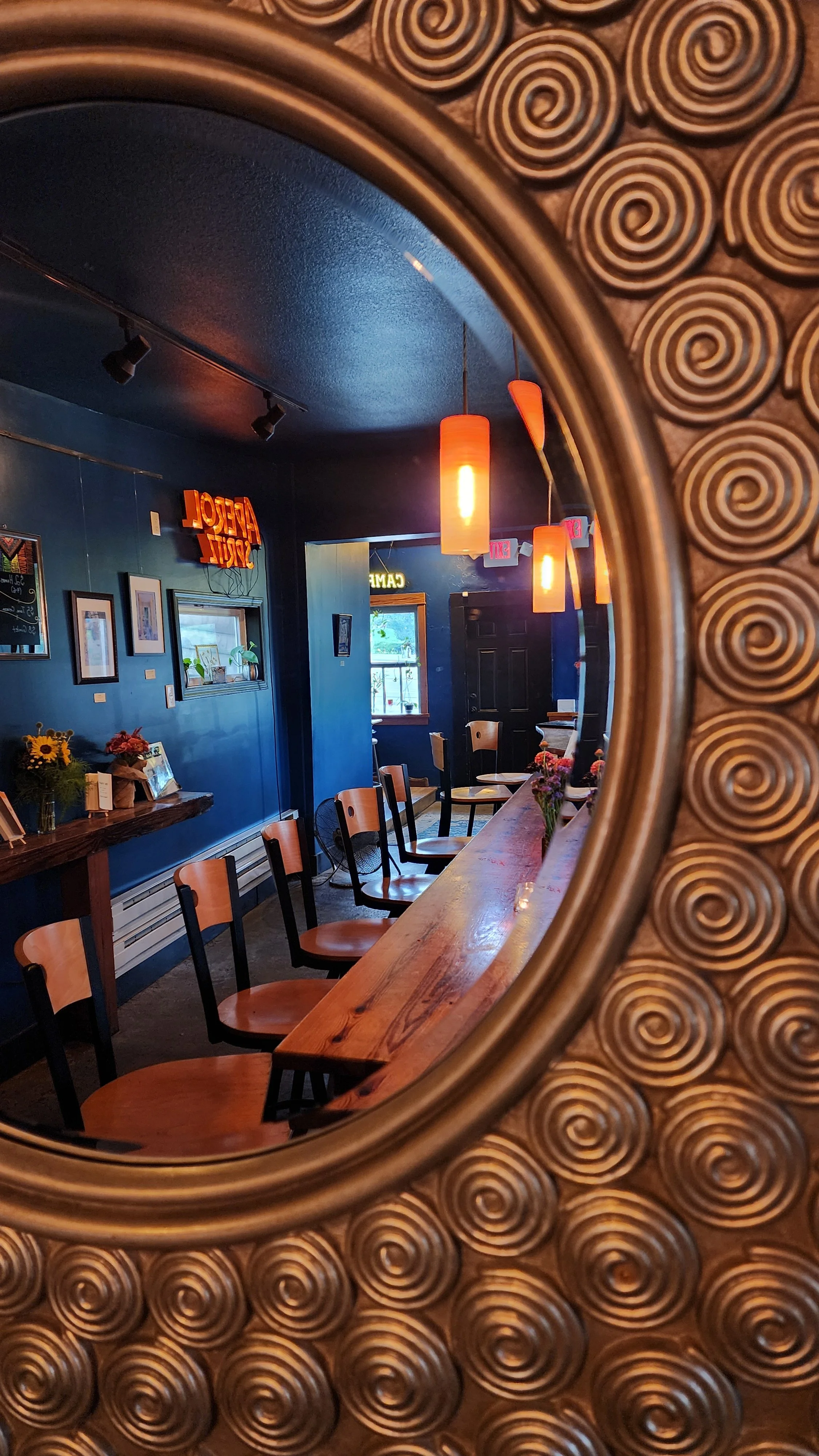 Interior of a cozy café, seen through a decorative circular mirror frame with spiral patterns. The café has wooden chairs, a long wooden bar, hanging orange pendant lights, framed pictures and plants on the wall, and a window with natural light.