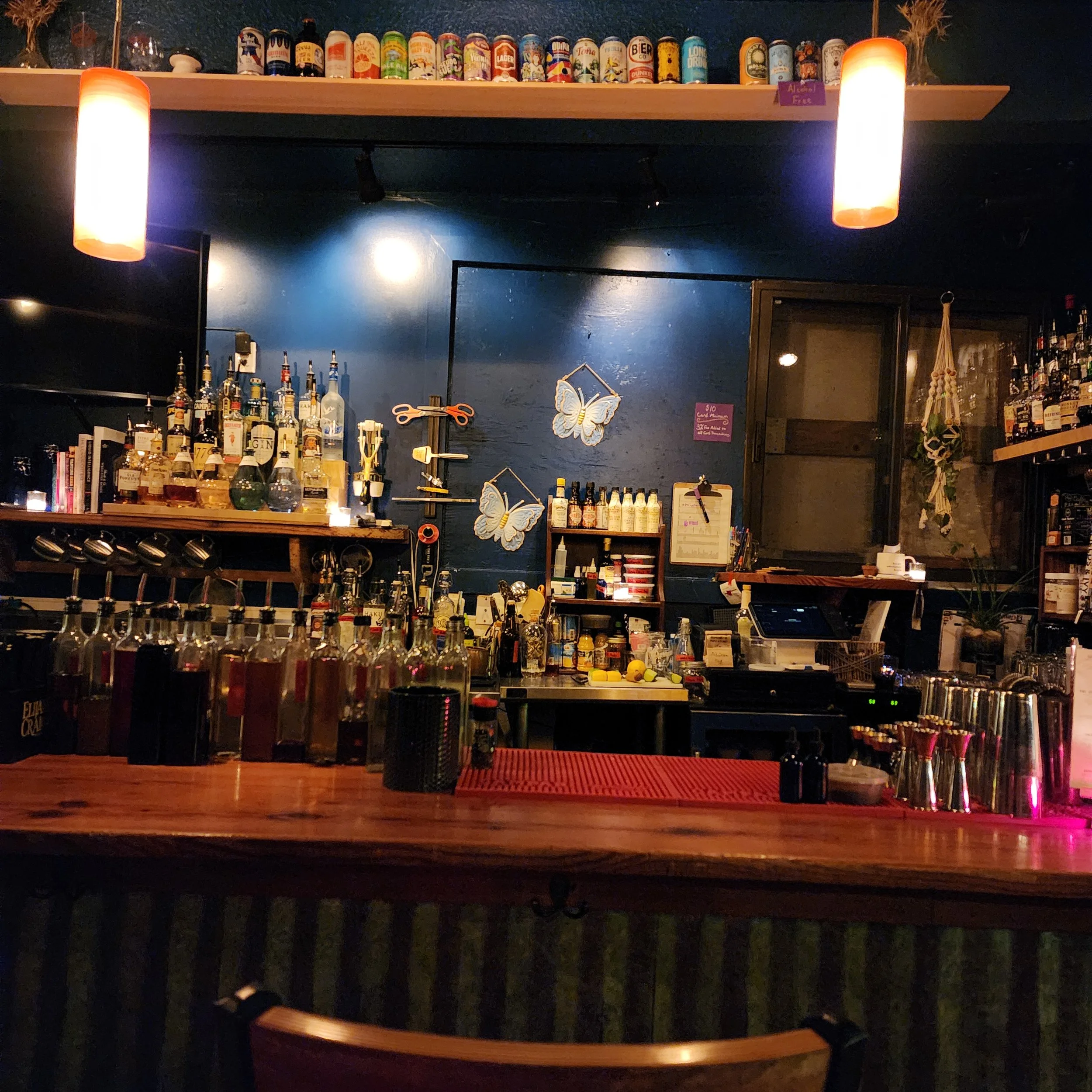 A bar with shelves of colorful cans and bottles, illuminated by warm orange hanging lights, with bottles and bar tools on the counter.