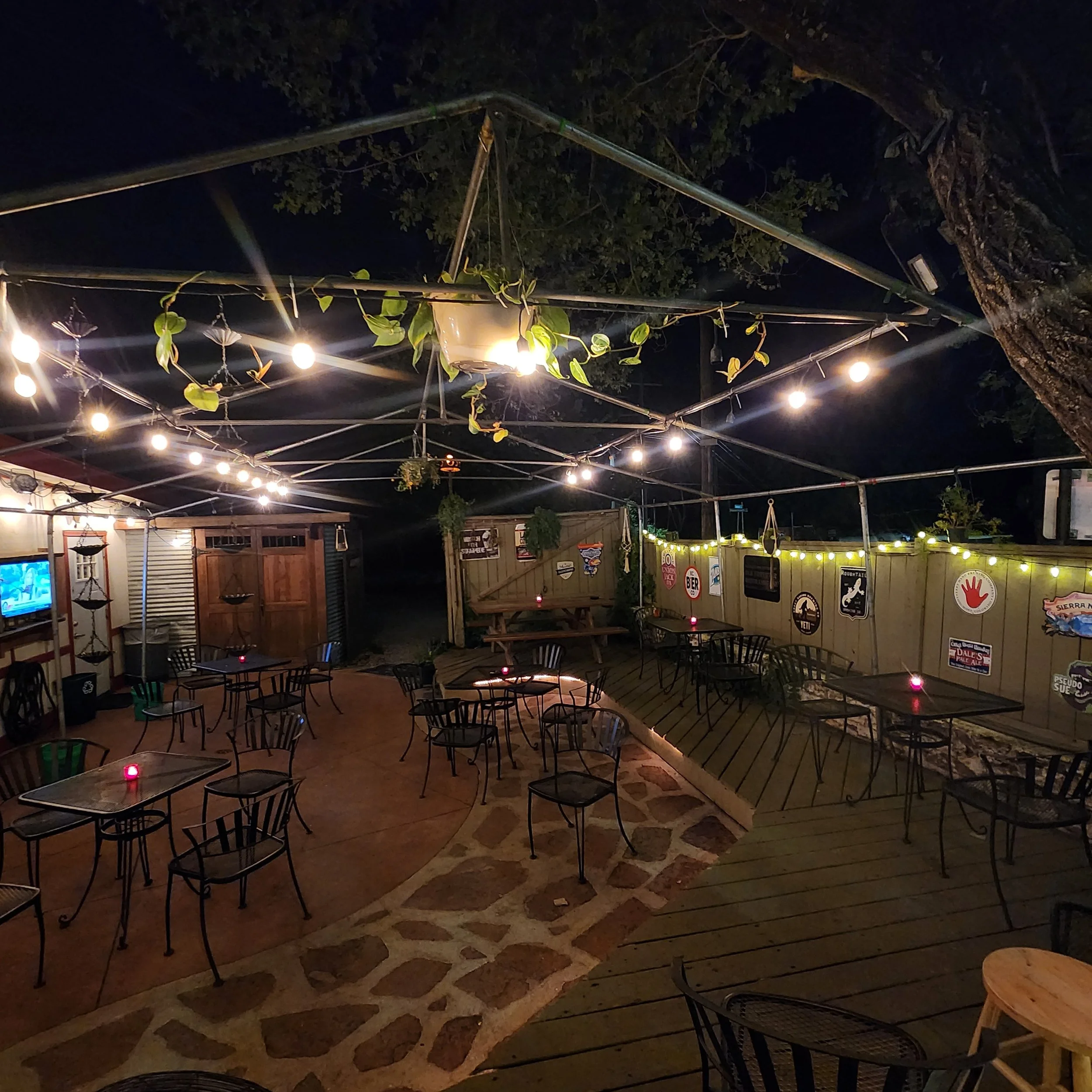 Outdoor patio area at night with string lights hanging from a metal frame and a small hanging planter with green vines. Wooden fence with various signs and a TV visible in the background. Tables and chairs set up on a stone-paved surface.