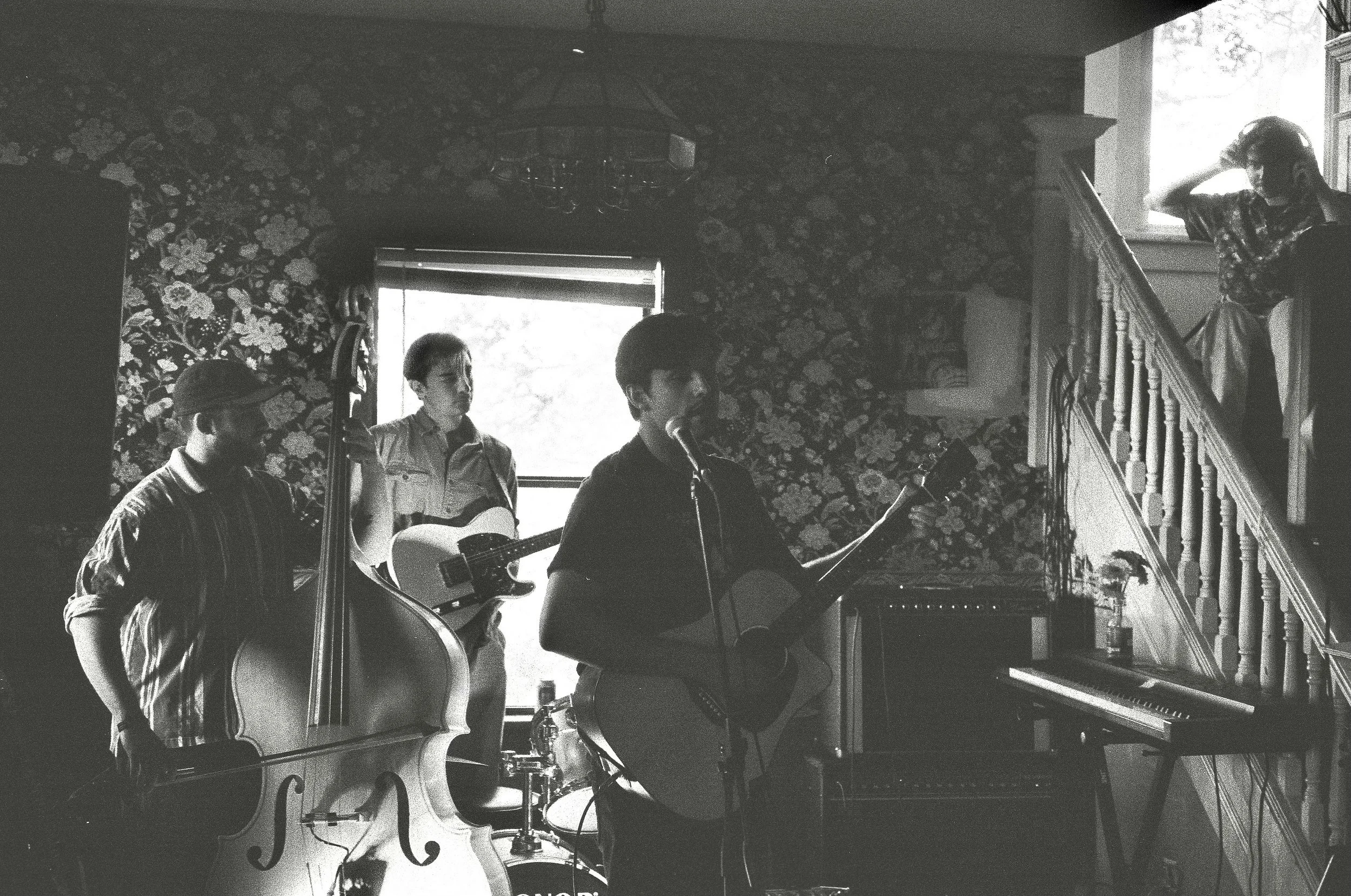 A black and white photo of a group of musicians performing indoors, with some on guitars and a microphone, against a floral-patterned wall with a window and staircase visible.