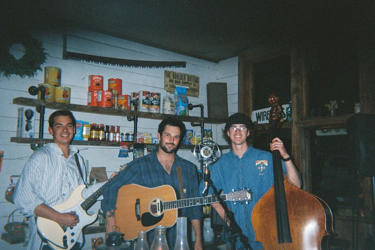 Three men holding guitars in a cozy, rustic decorated room with shelves of canned goods and a sign that reads "The Boulder Bouting and Bar Supply."