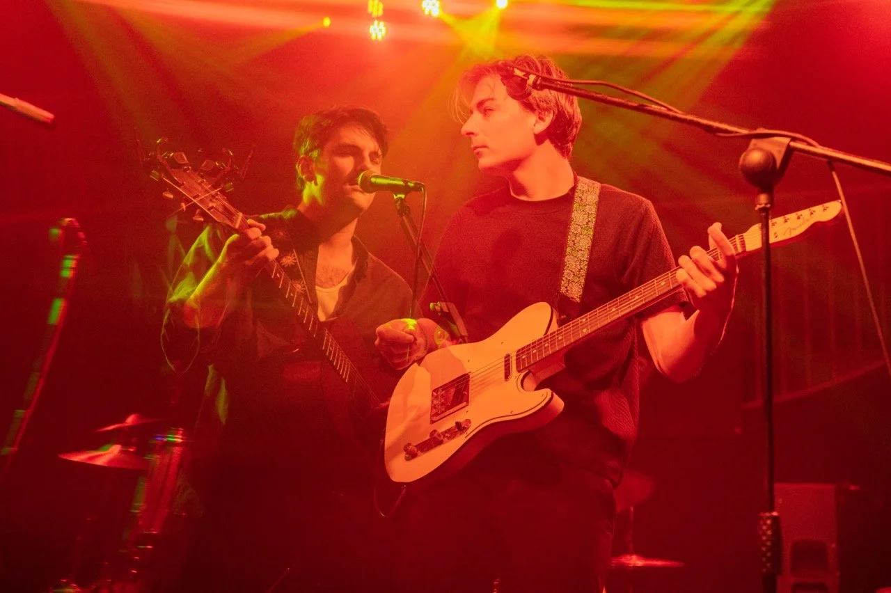 Two male musicians playing guitars and singing on stage under red and yellow lights.