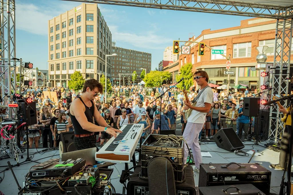 Outdoor concert on a city street with a band performing on stage and a crowd watching. Urban buildings and traffic lights are visible in the background.