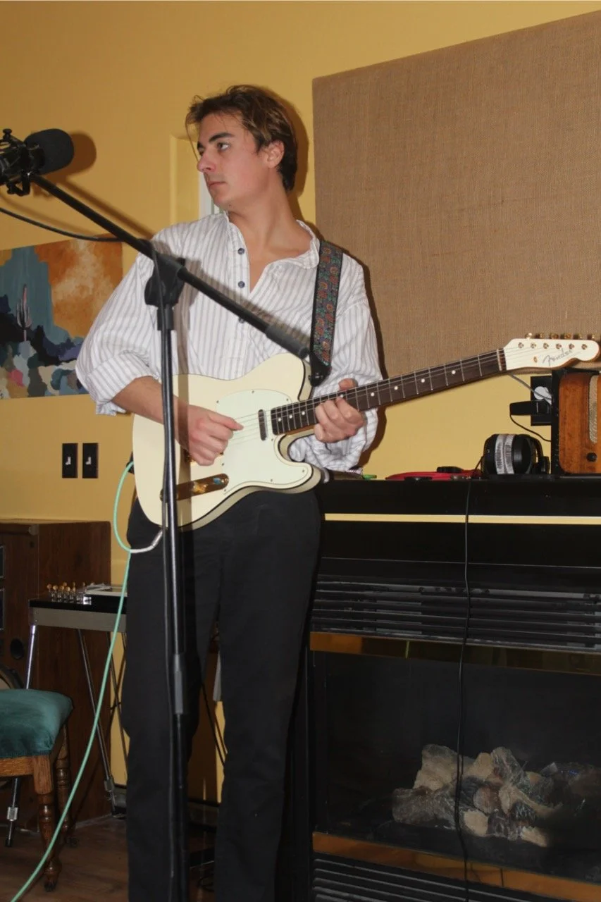 Young man with brown hair playing a white electric guitar indoors, surrounded by music equipment.