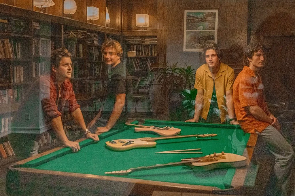 Four young men in casual clothing standing and sitting around a green pool table with musical instruments, in a room with wood-paneled walls and bookshelves.
