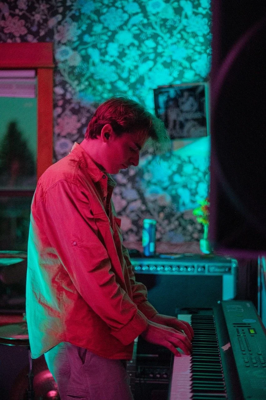 A person playing a keyboard in a dimly lit room with colorful lighting and a stone wall background.