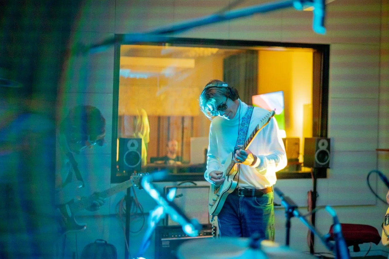 A person with headphones and glasses playing an electric guitar in a music studio, with another person in the background working on music equipment.