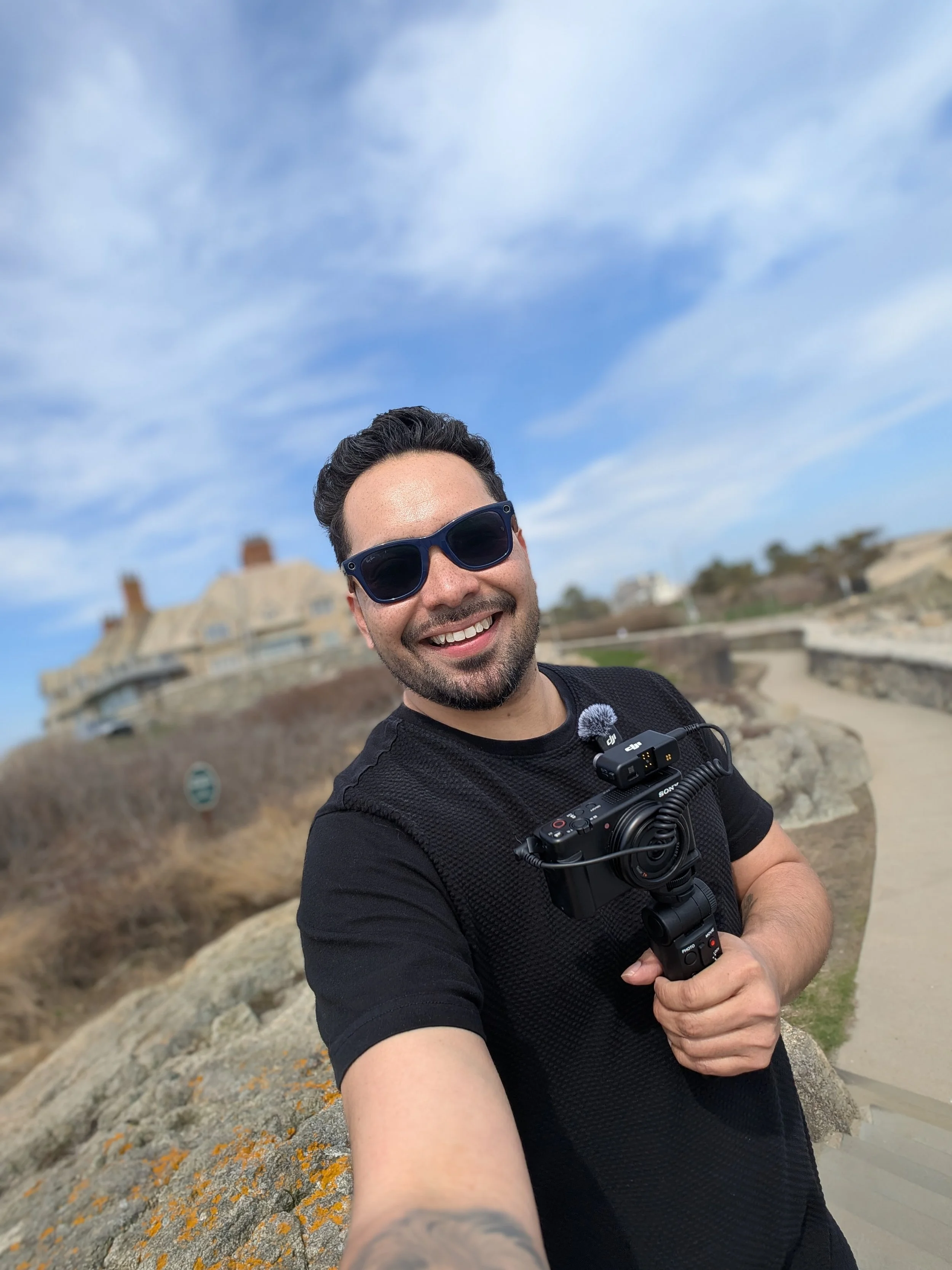 Hombre sonriendo, con gafas de sol y una cámara en mano, en un lugar con rocas y edificios al fondo bajo un cielo azul con algunas nubes.